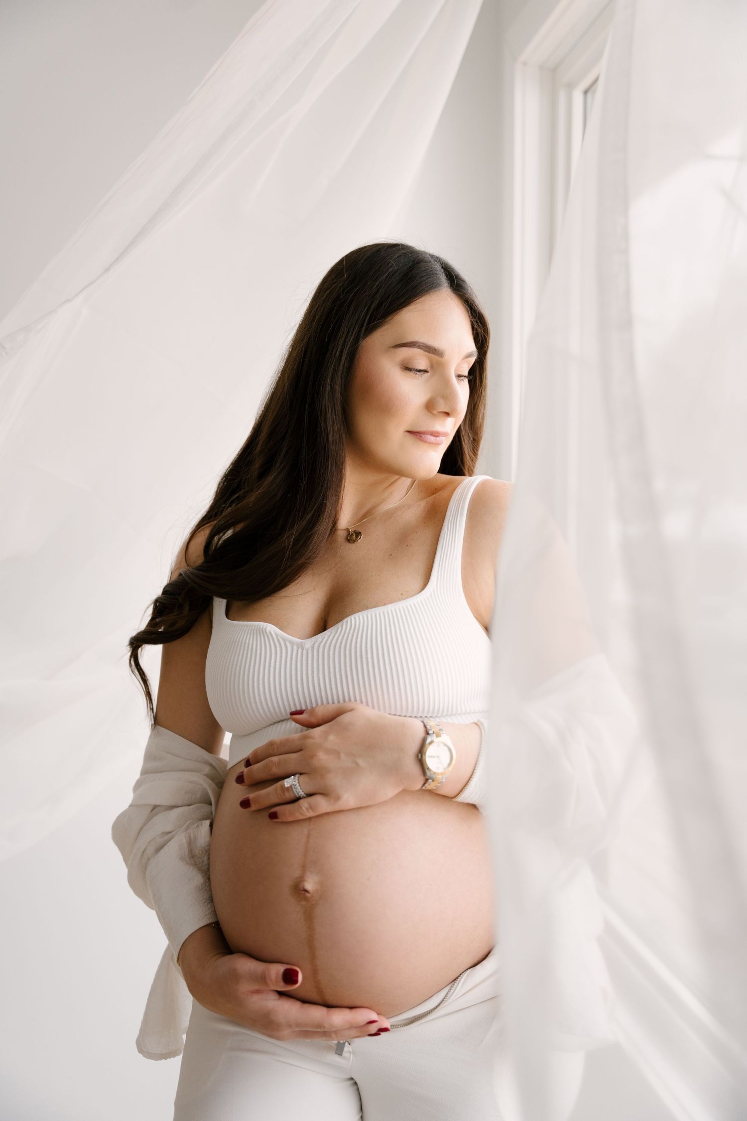Beautiful pregnant woman in white crop top holding baby bump near sheer curtains in bright natural light.
