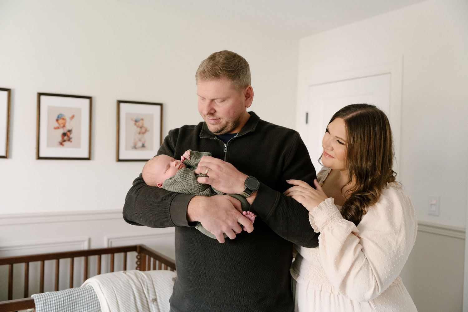 New parents admire their newborn baby in a beautifully decorated nursery with framed art on the wall.