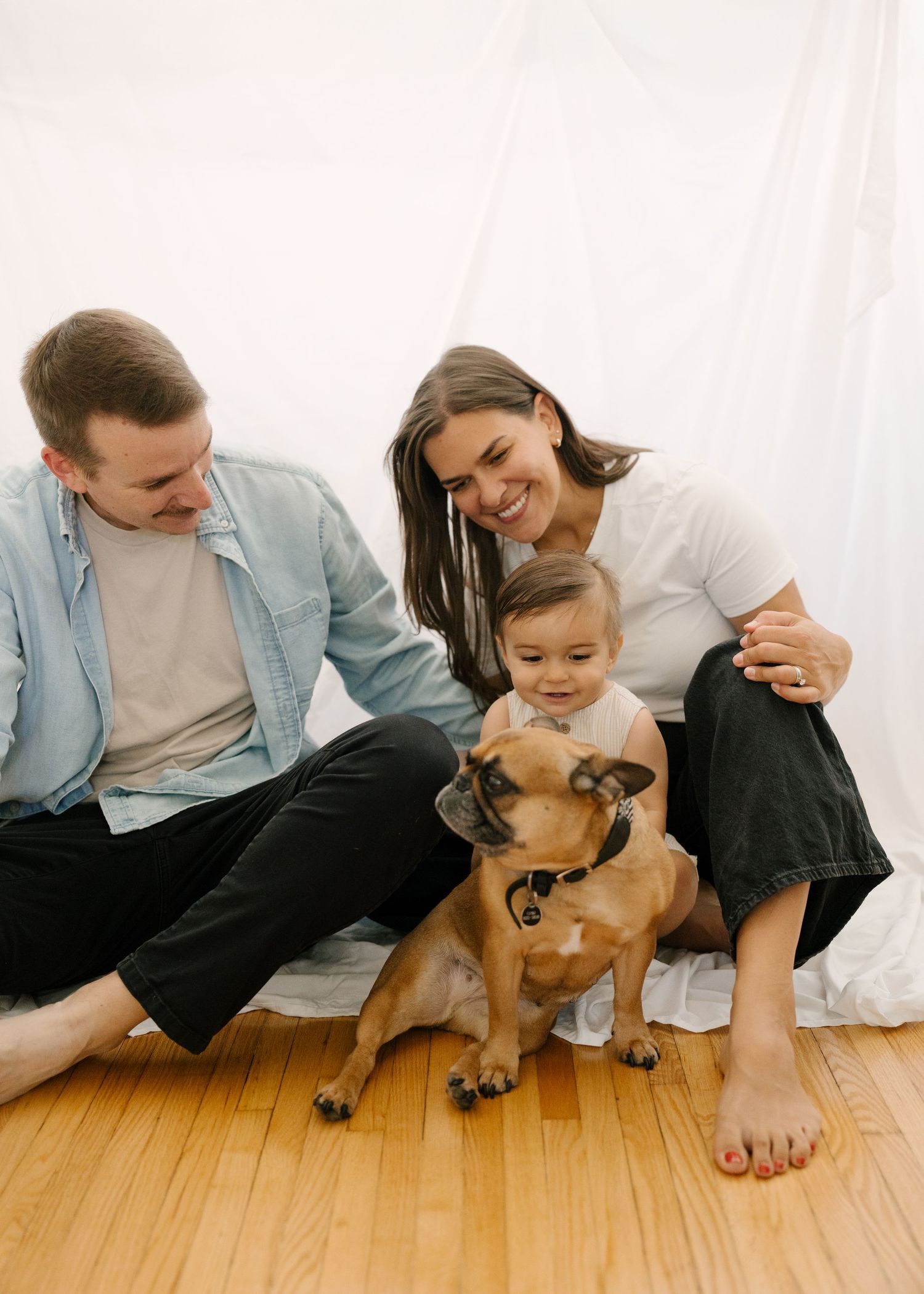 Happy couple with their baby and brown dog sit together on a hardwood floor with white backdrop.