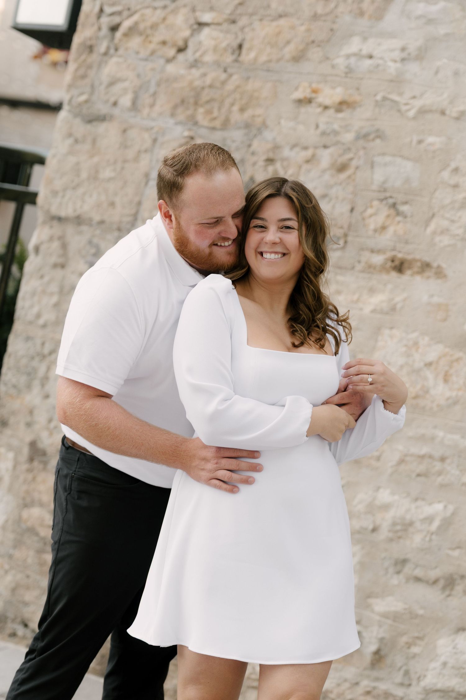 Smiling couple embracing in front of a rustic stone wall, woman wearing a white mini dress.