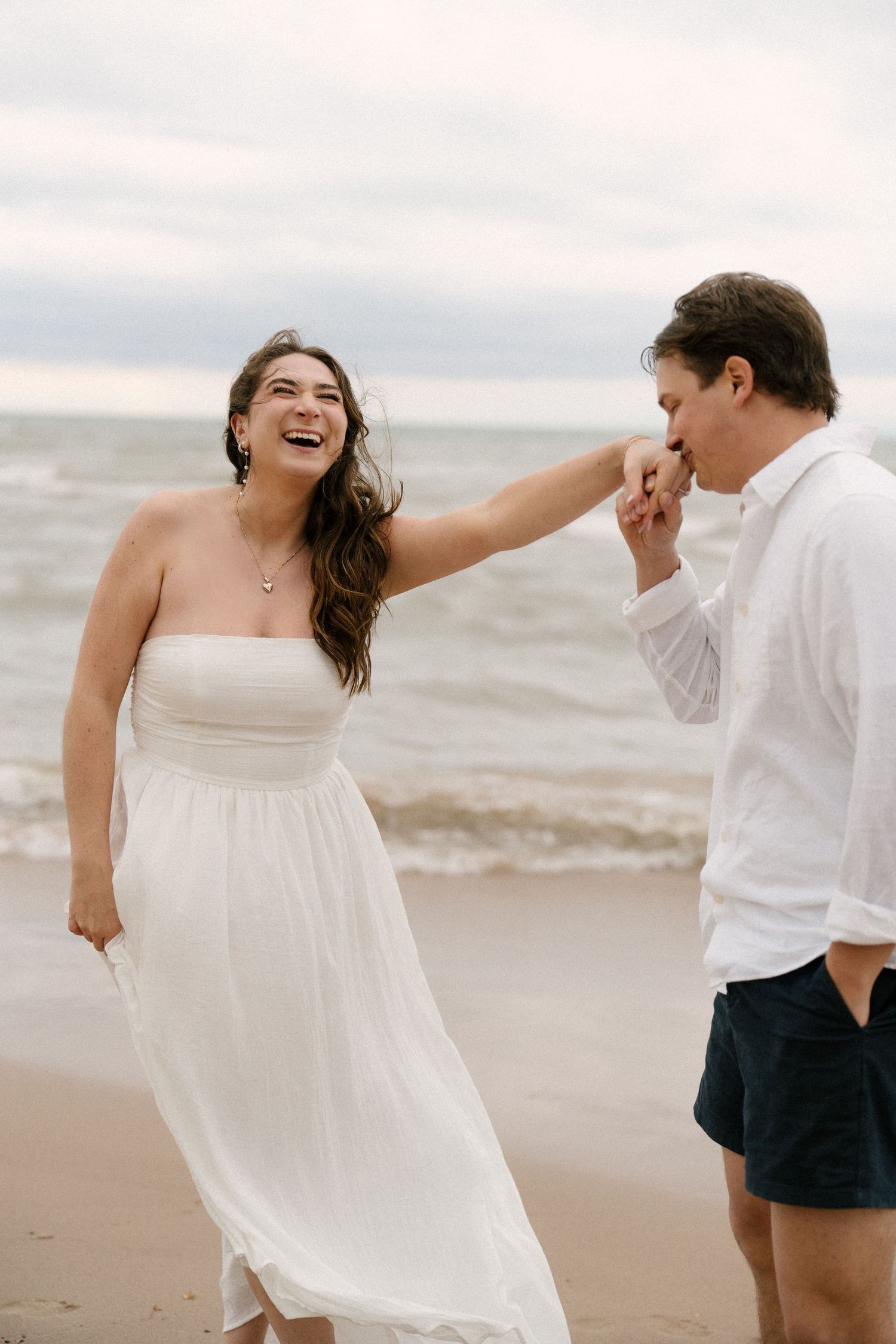 Woman in white strapless dress laughing joyfully as man kisses her hand on a sandy beach by the ocean.