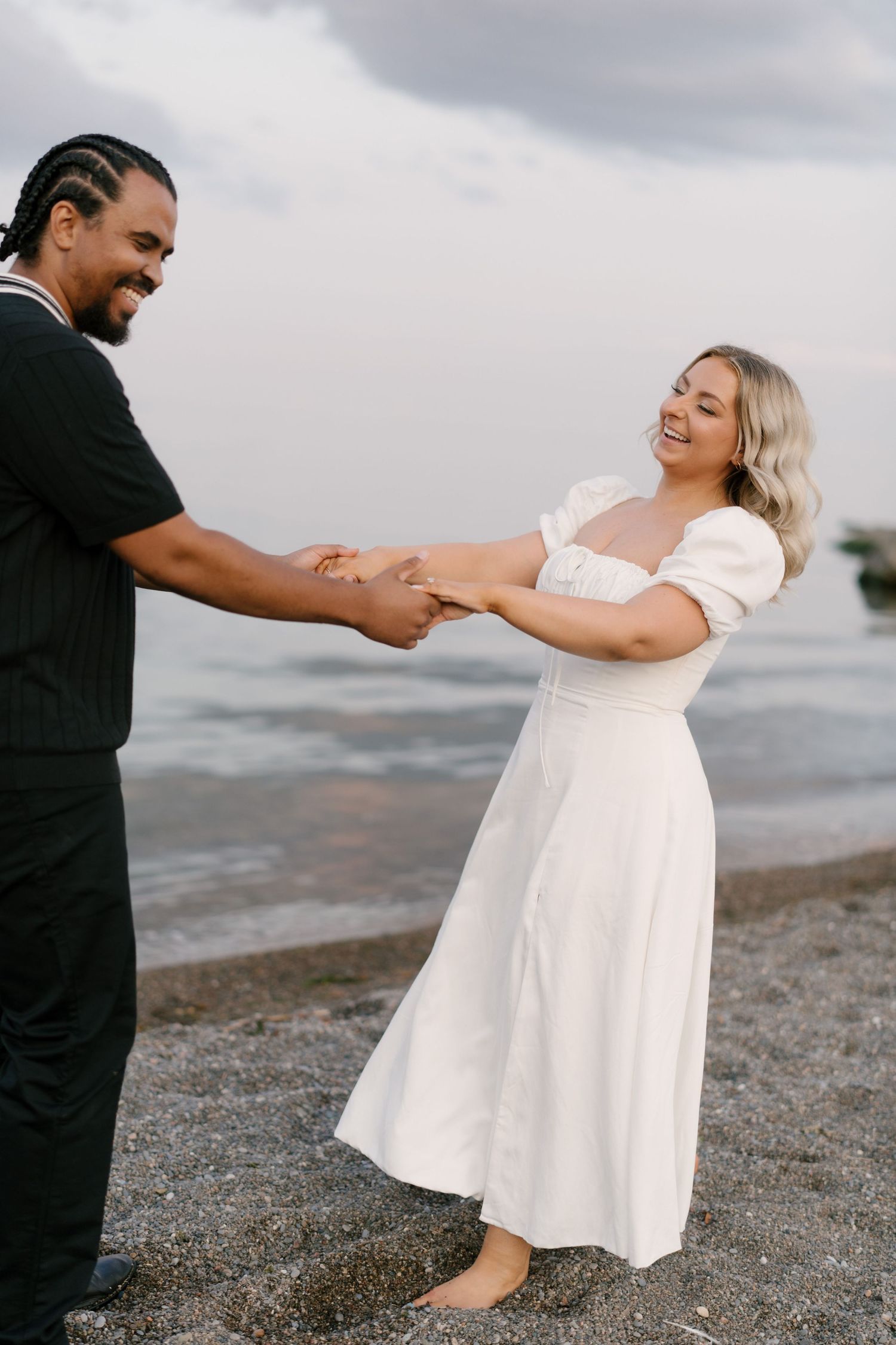 A joyful couple laughs and holds hands together on a pebble beach with ocean waves in the background.