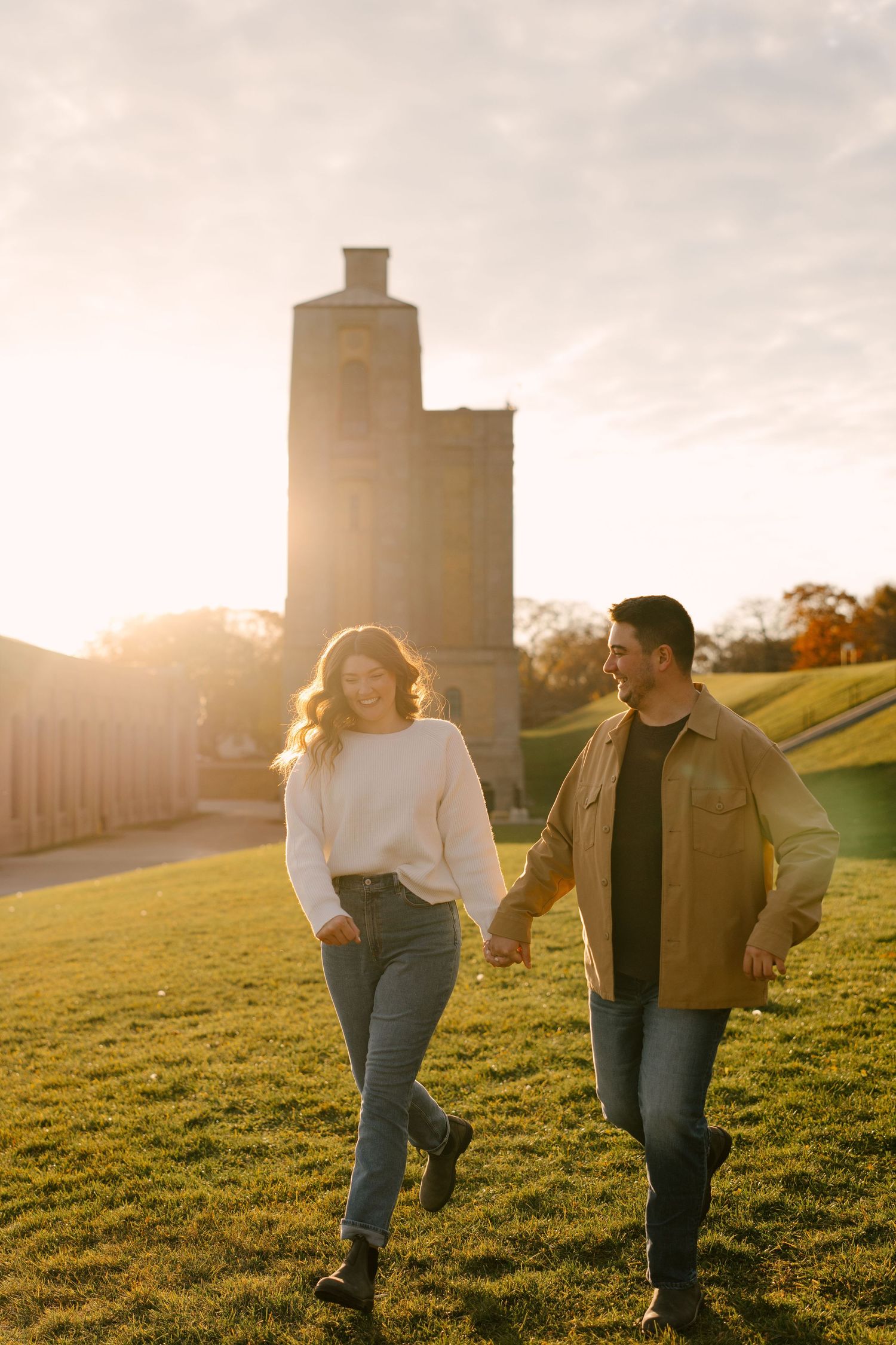 Couple holding hands walking on green grass at golden hour with historic stone tower in background.