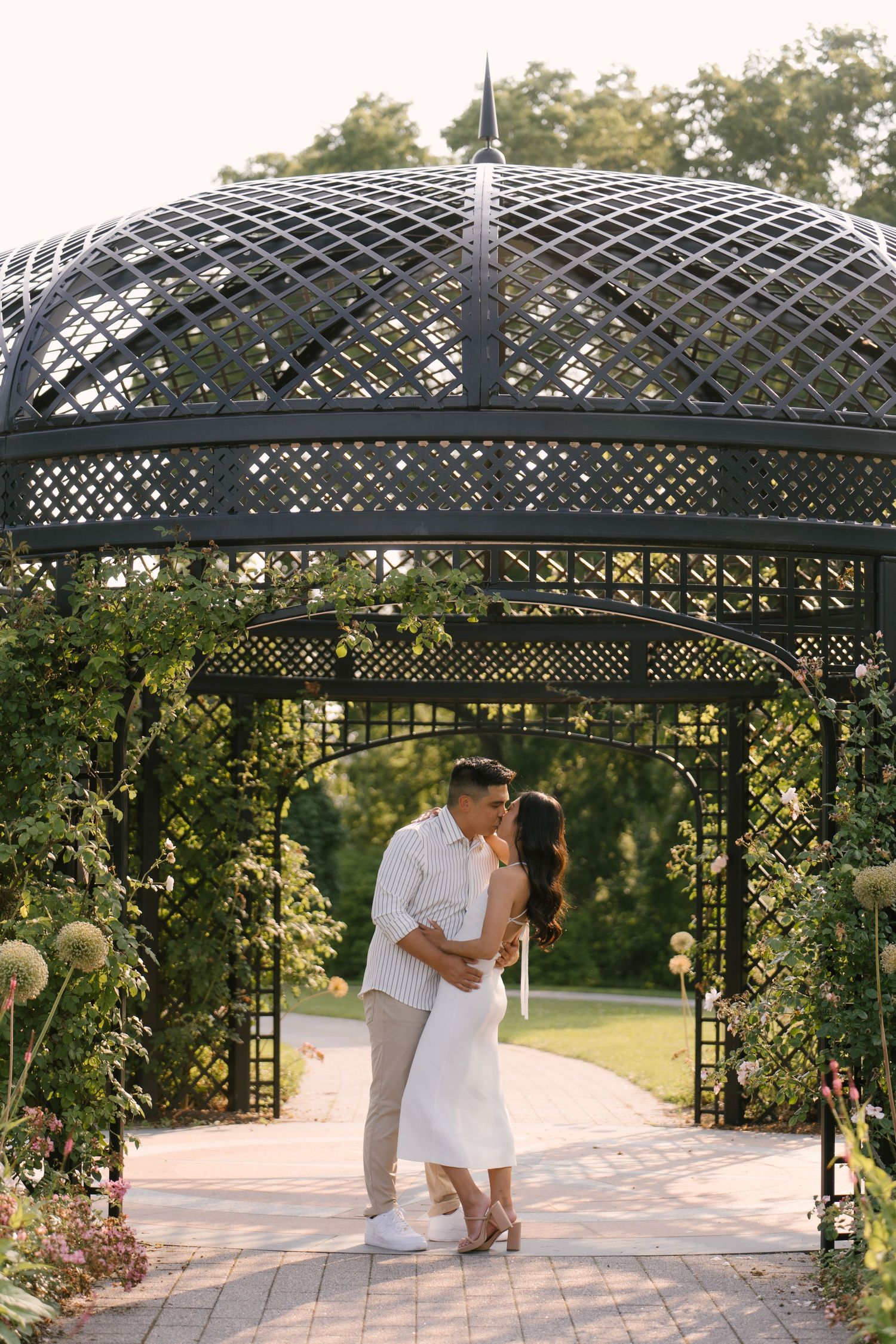 Couple sharing a romantic embrace under an elegant black lattice gazebo surrounded by lush garden flowers.