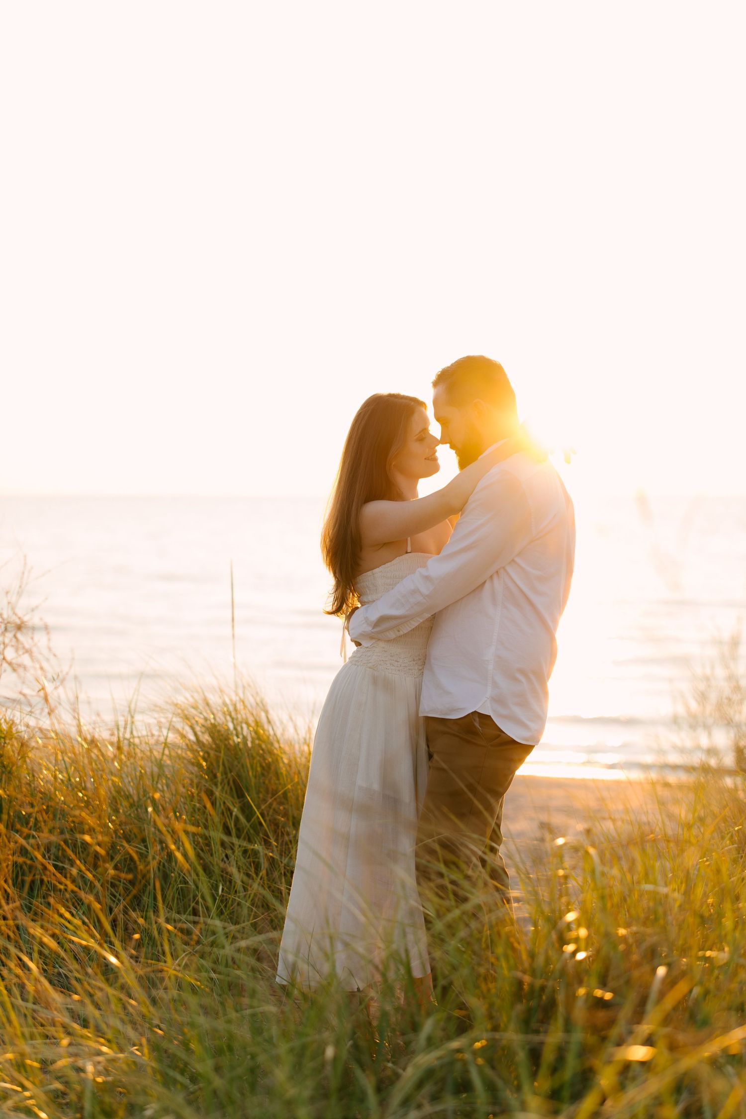 Couple embracing in golden sunset light among beach grass by the ocean.