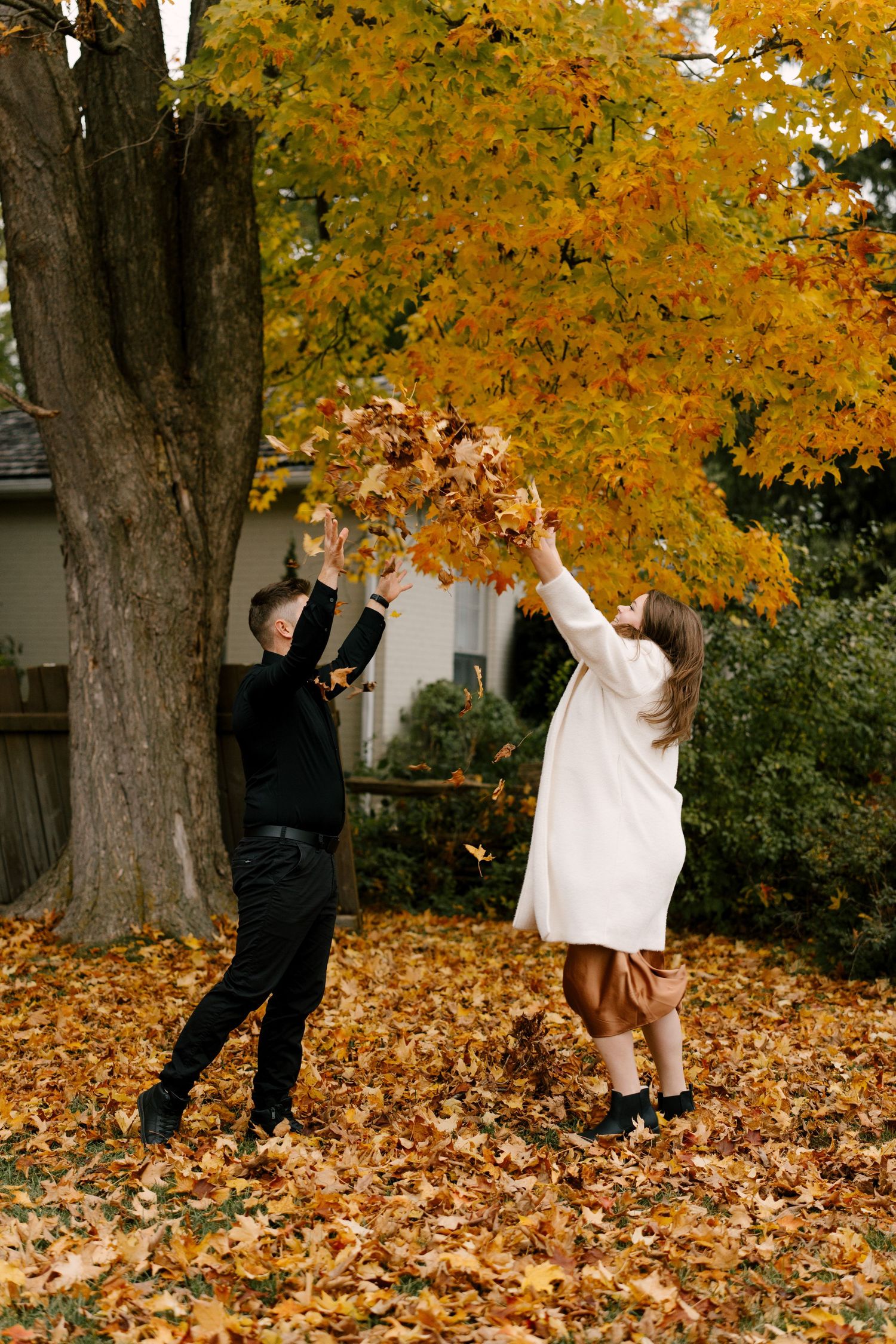 Joyful couple tossing golden autumn leaves under a vibrant yellow maple tree during a fall photo shoot.
