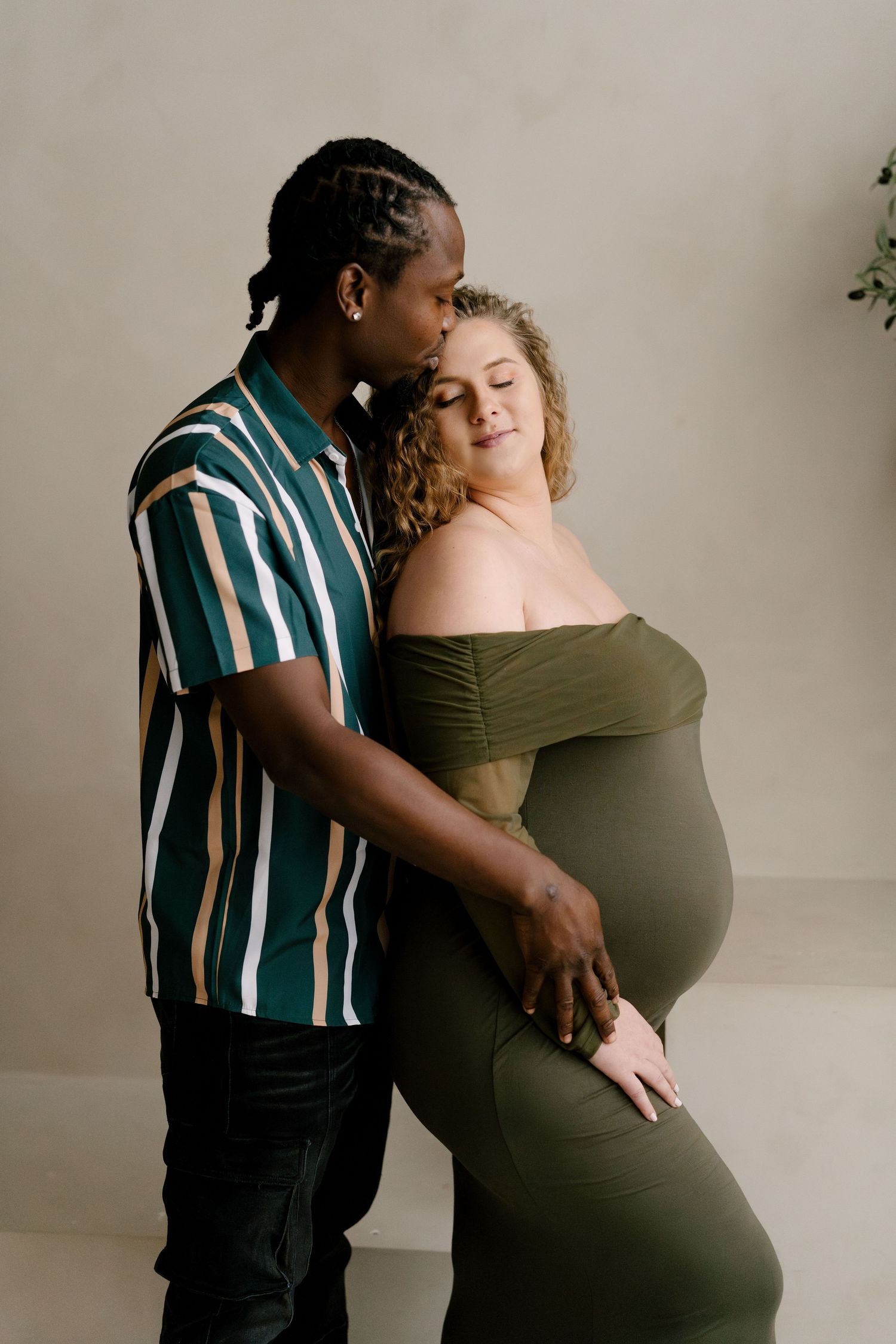 Interracial couple maternity photo, man kissing pregnant woman in olive off-shoulder gown in studio.
