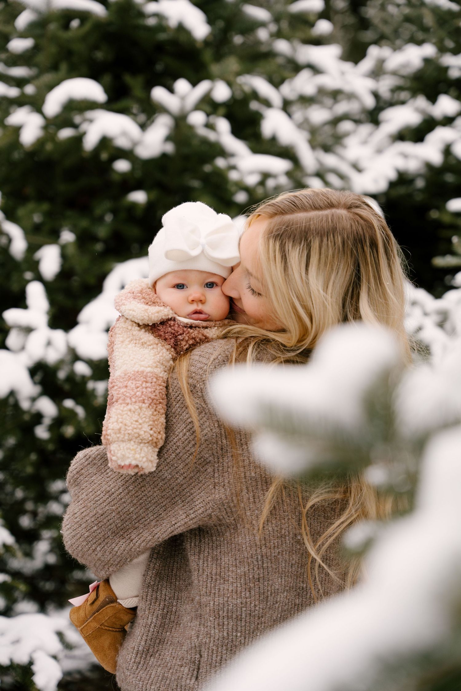 Blonde mother kissing her baby girl wearing white bow hat among snow-covered evergreen trees.