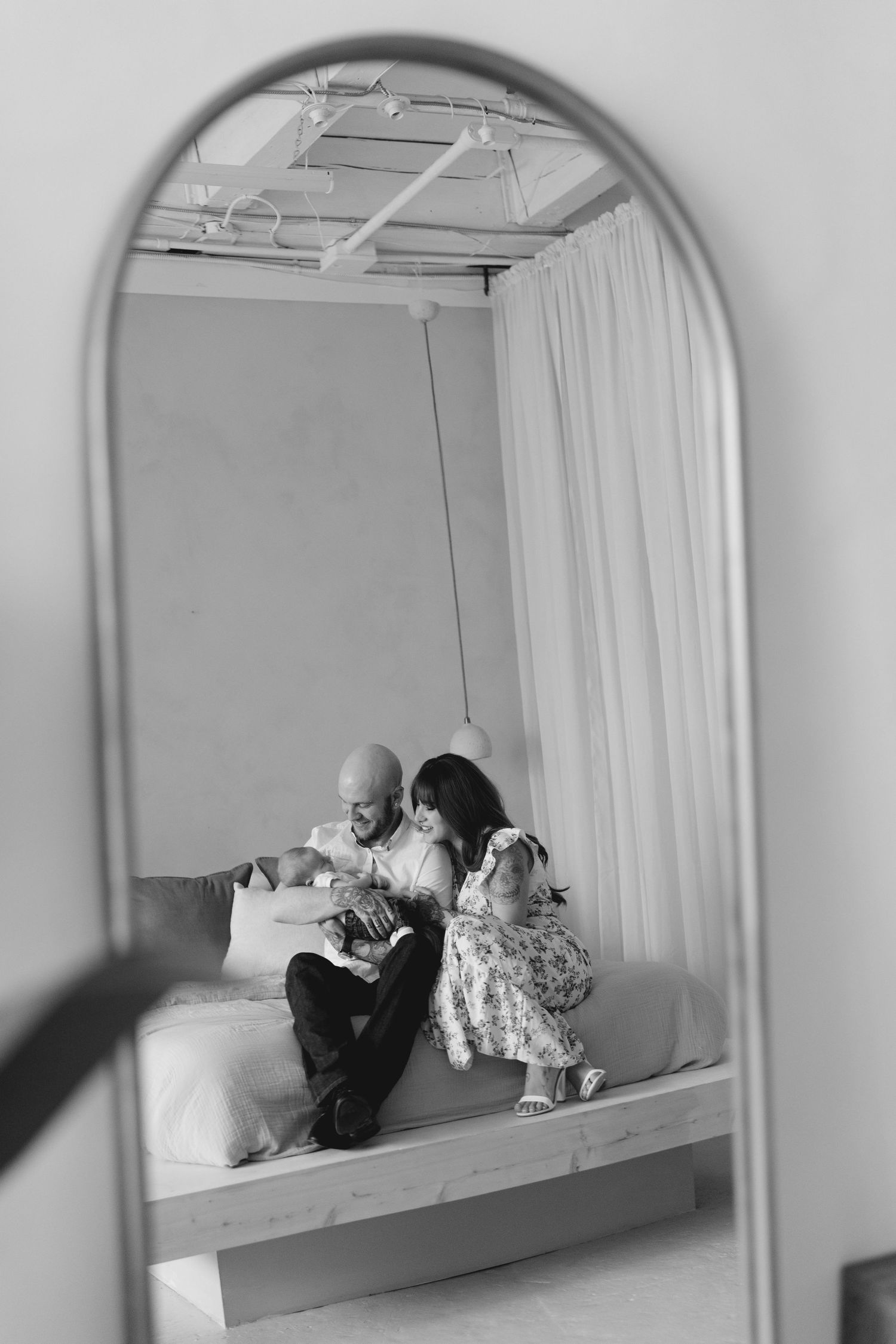 Black and white mirror reflection of a couple sitting on a bed admiring their newborn baby.