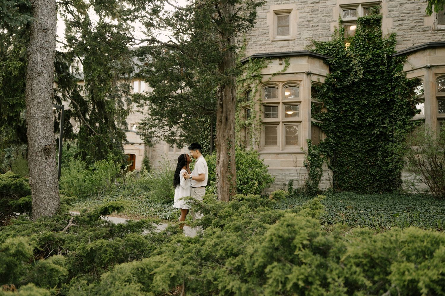 Couple embracing among green shrubs in a garden next to an ivy-covered building.