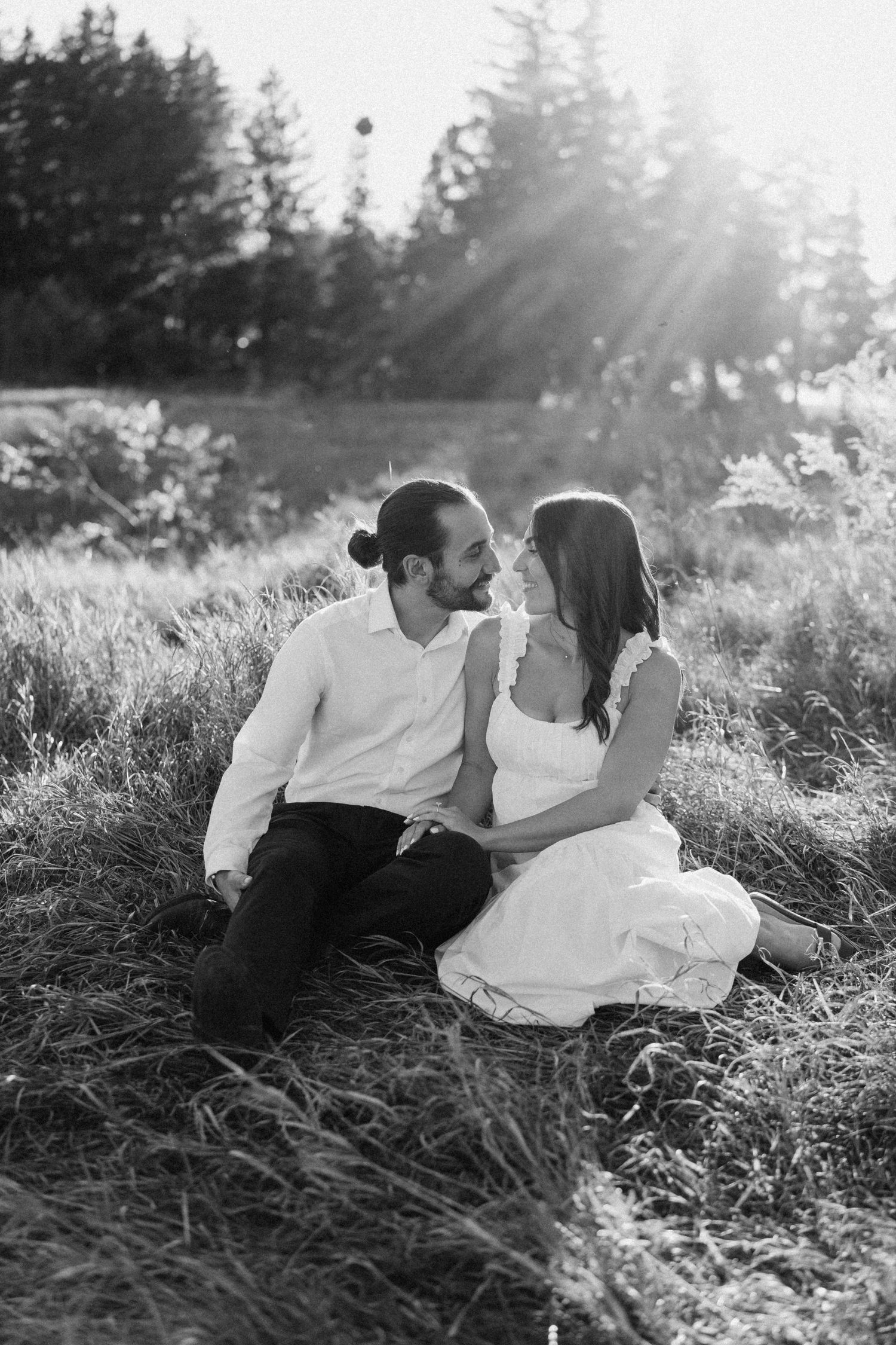 Black and white photo of a couple sitting in a sunlit meadow, gazing at each other romantically.