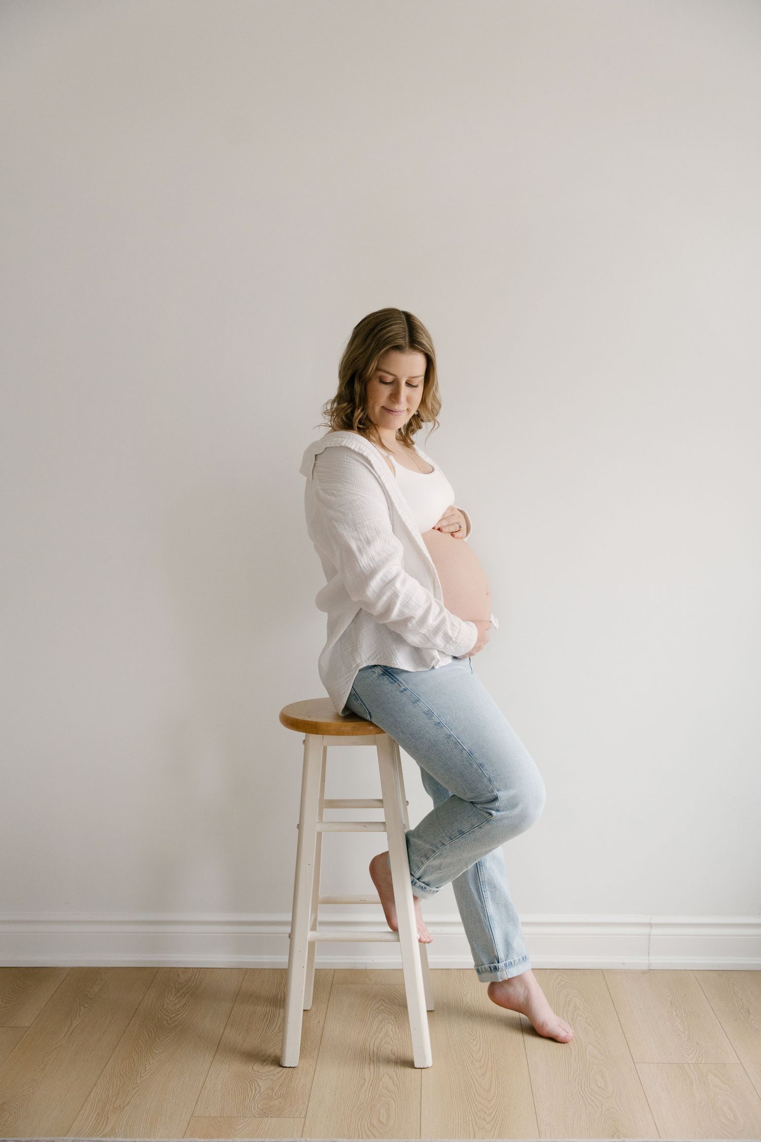 Pregnant woman in white jacket and jeans sitting on a stool, cradling her baby bump indoors.