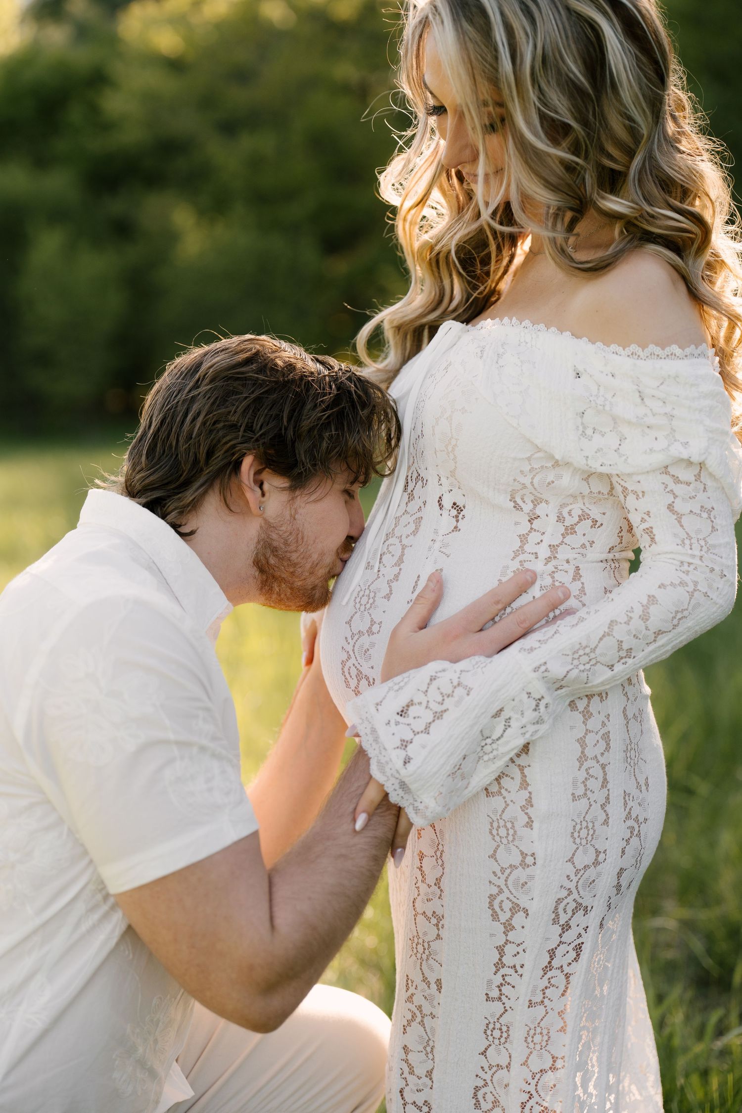 Man kneeling and kissing pregnant woman's belly outdoors in a sunny green field during maternity shoot.