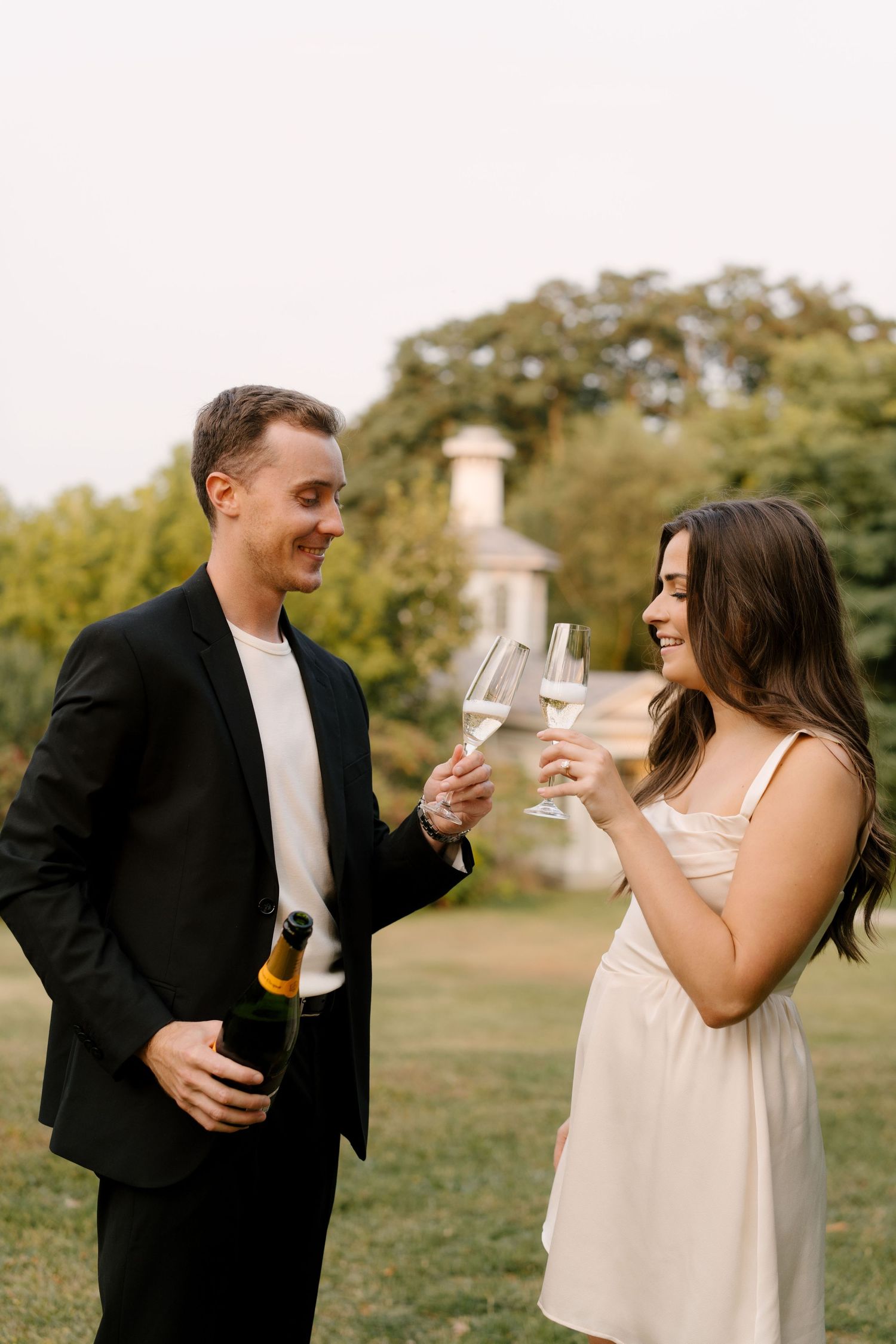 Joyful couple toasting champagne glasses outdoors near historic gazebo surrounded by lush green trees.