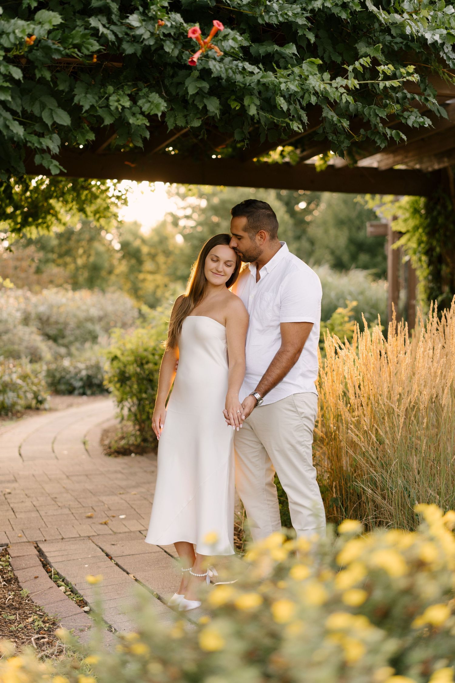 Man embracing woman in white strapless dress under vine-covered pergola in sun-drenched garden setting.