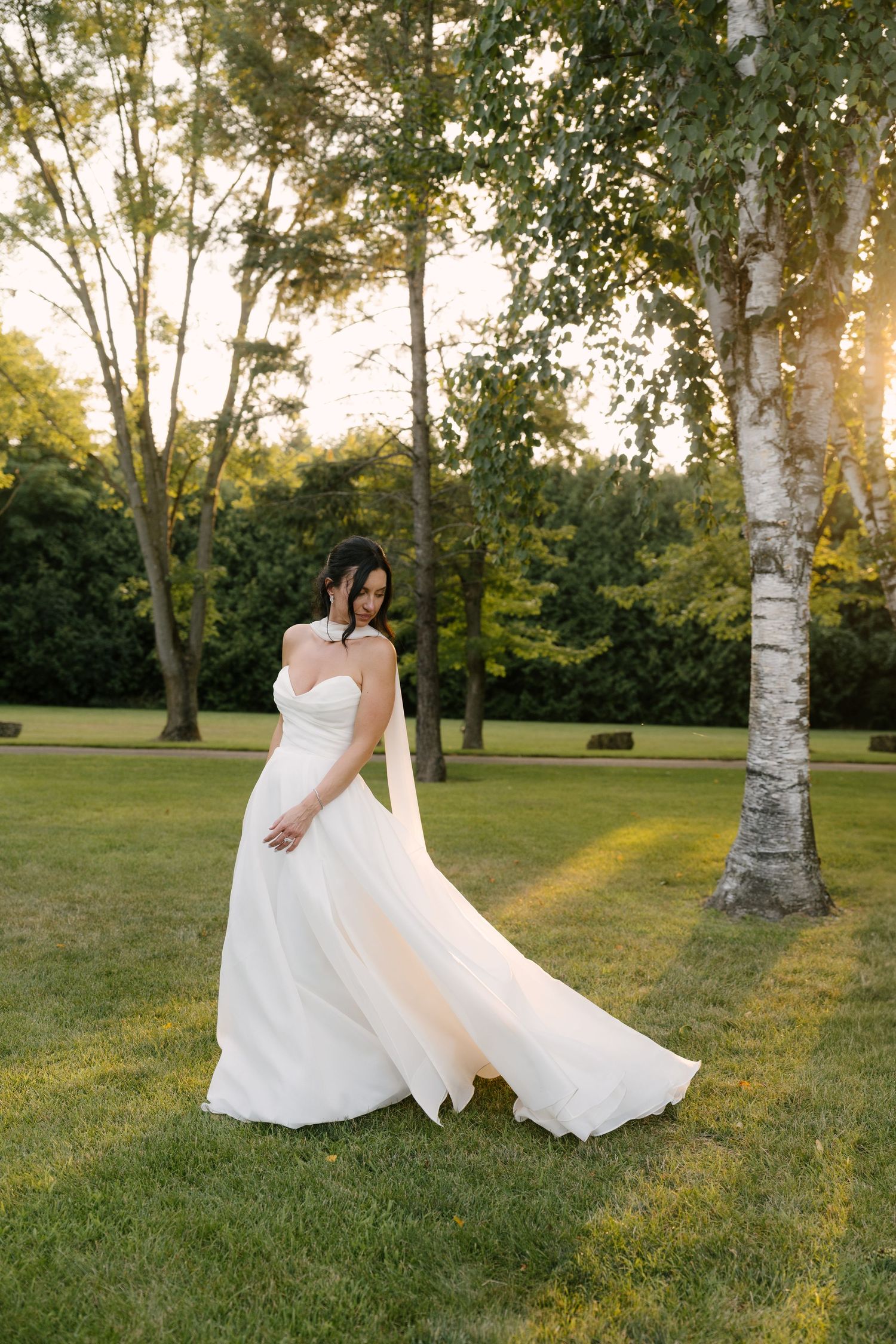 Bride in flowing white strapless gown twirling in lush green garden at golden hour sunset at Roseville Estates wedding venue.