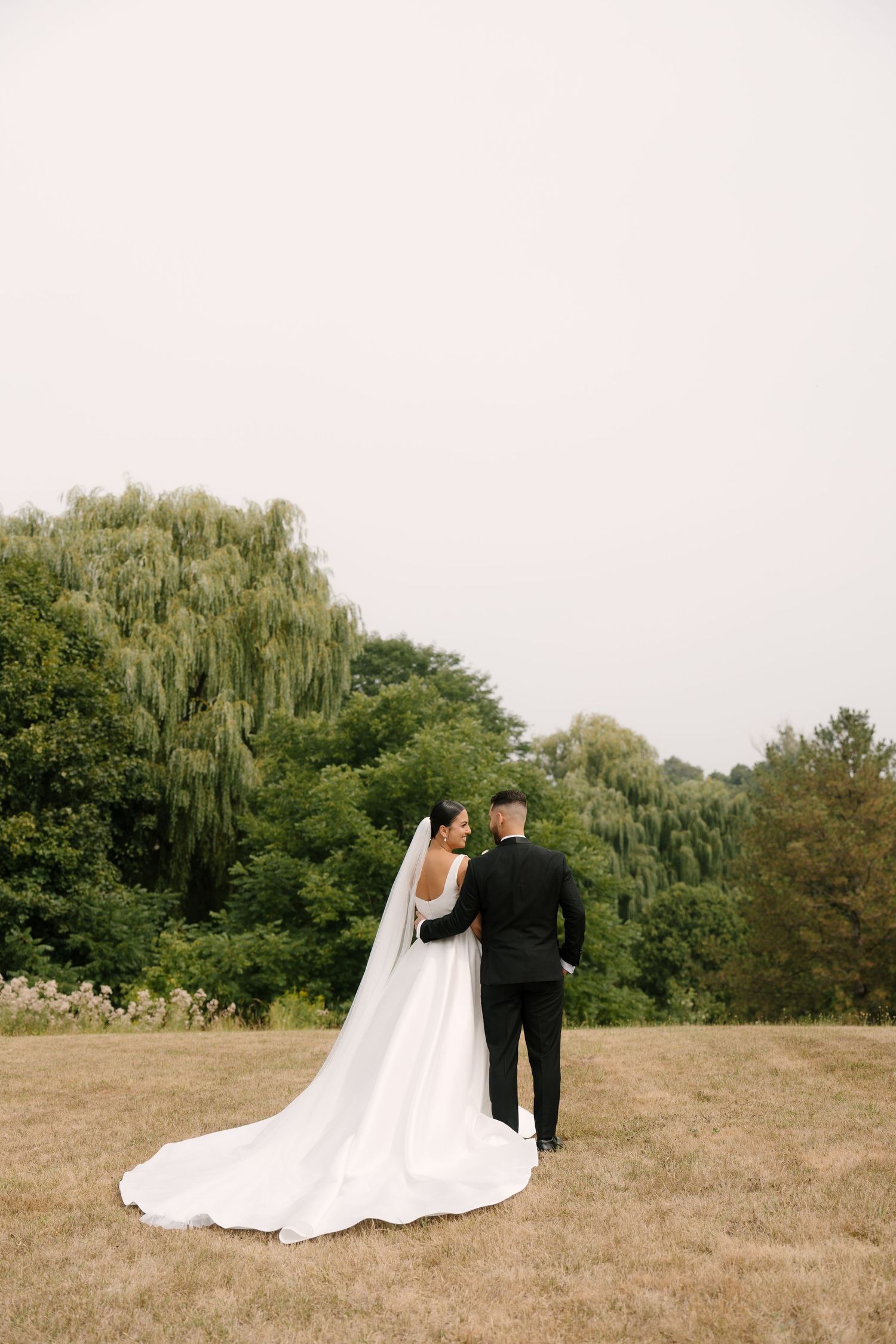 Bride and groom standing together in an open field, viewed from behind, with lush green trees in the background at parkwood Estates.