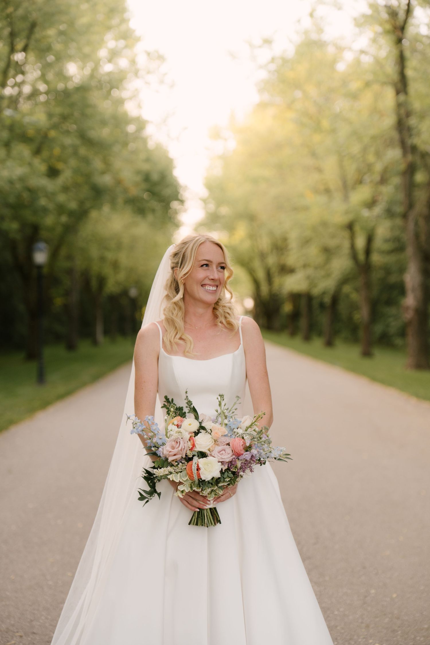 Smiling bride holding a pastel bouquet of pink and blue flowers on a sunlit tree-lined path at Roseville Estates venue.