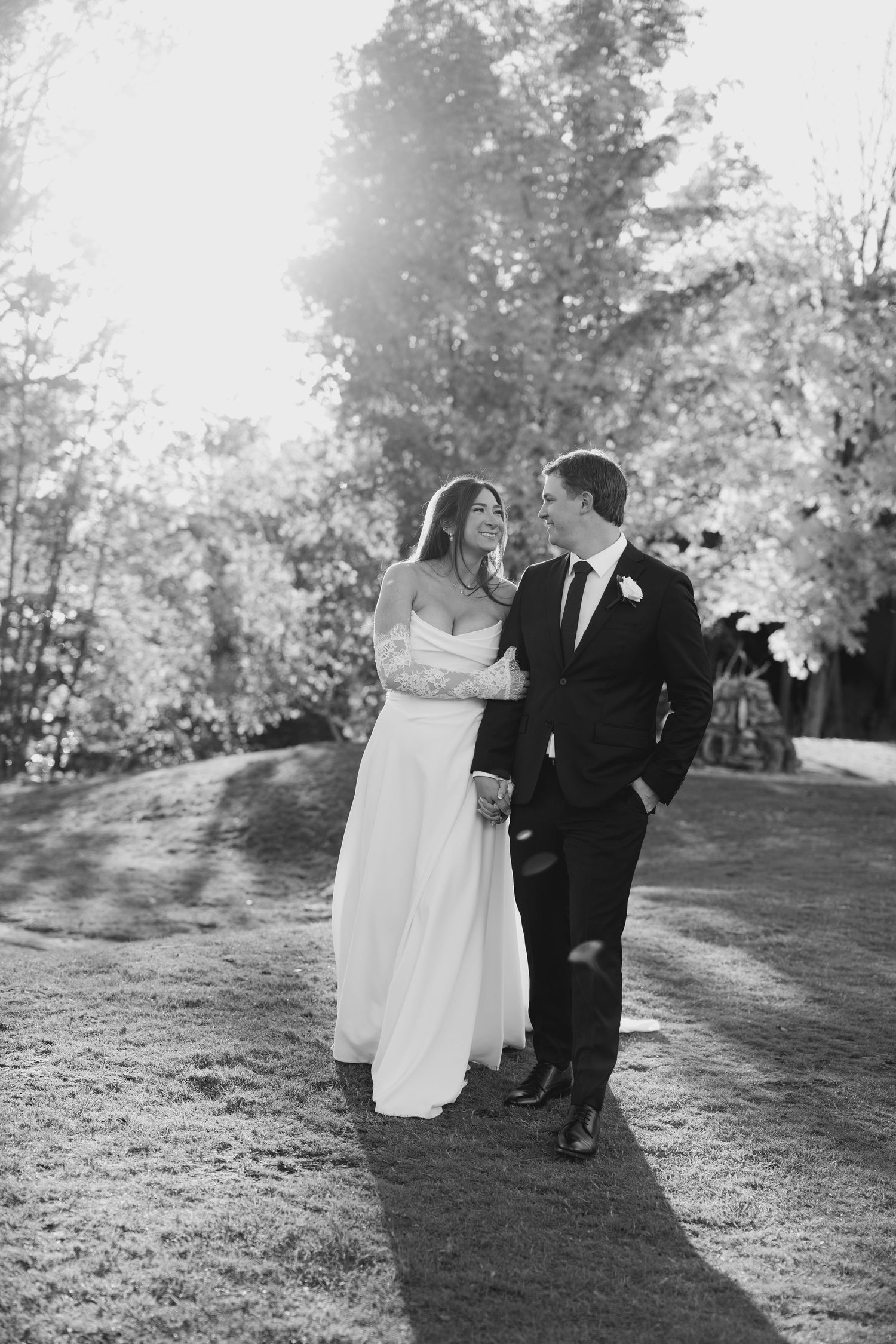 Bride and groom walk together on sunlit path through autumn trees, black and white wedding photo.