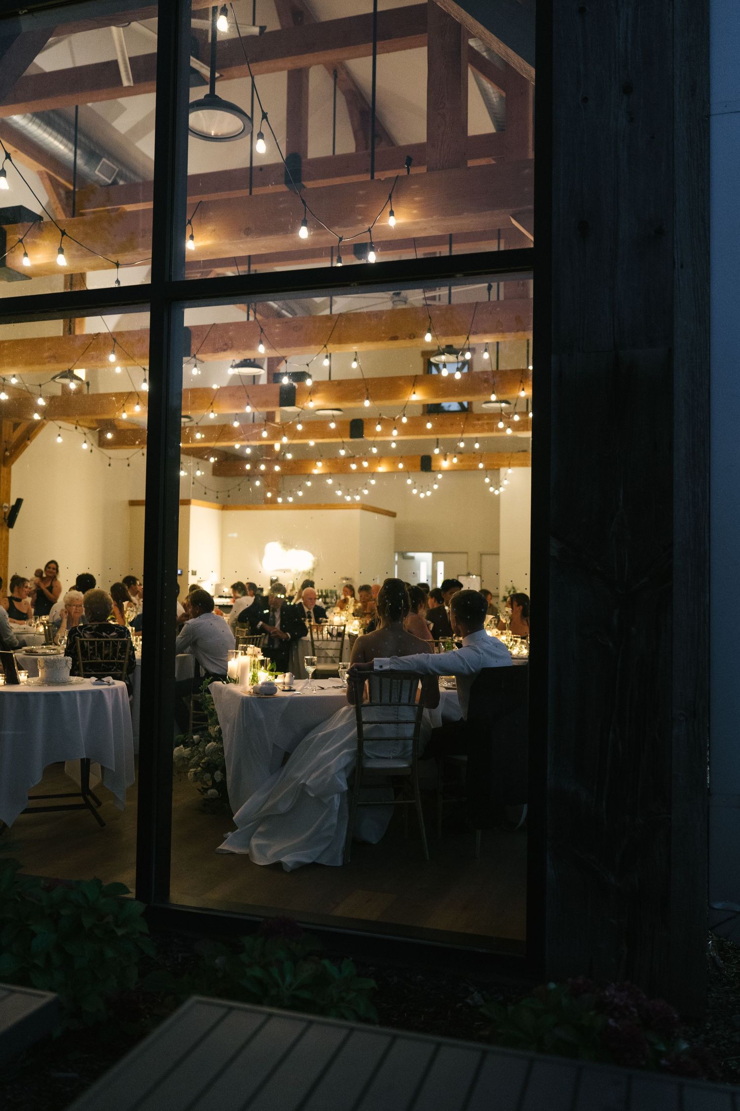 Elegant wedding reception viewed through large windows at night, featuring string lights and guests dining at candlelit tables.
