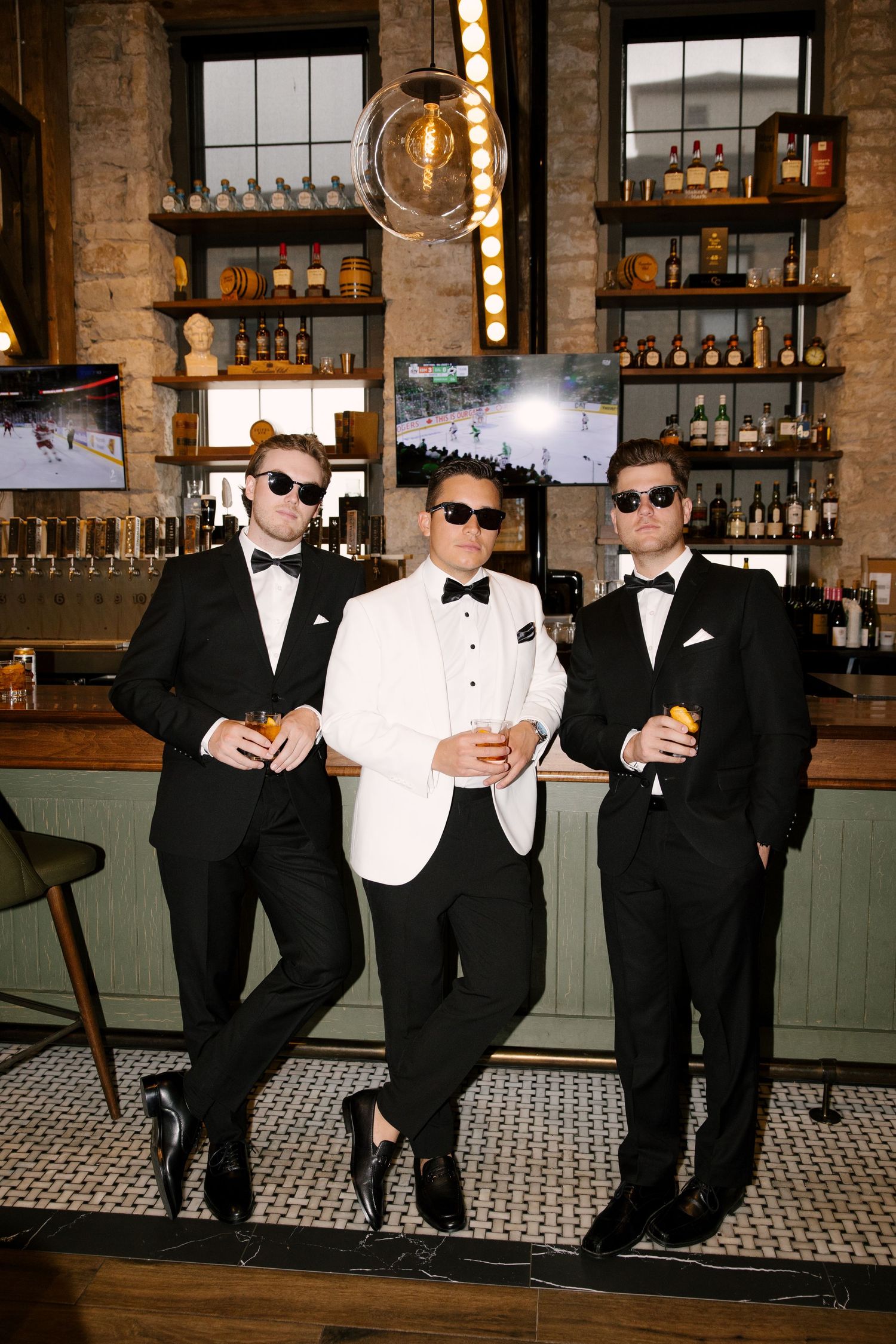 Groom in white tuxedo poses with two groomsmen in black suits holding drinks at The Tavern bar.