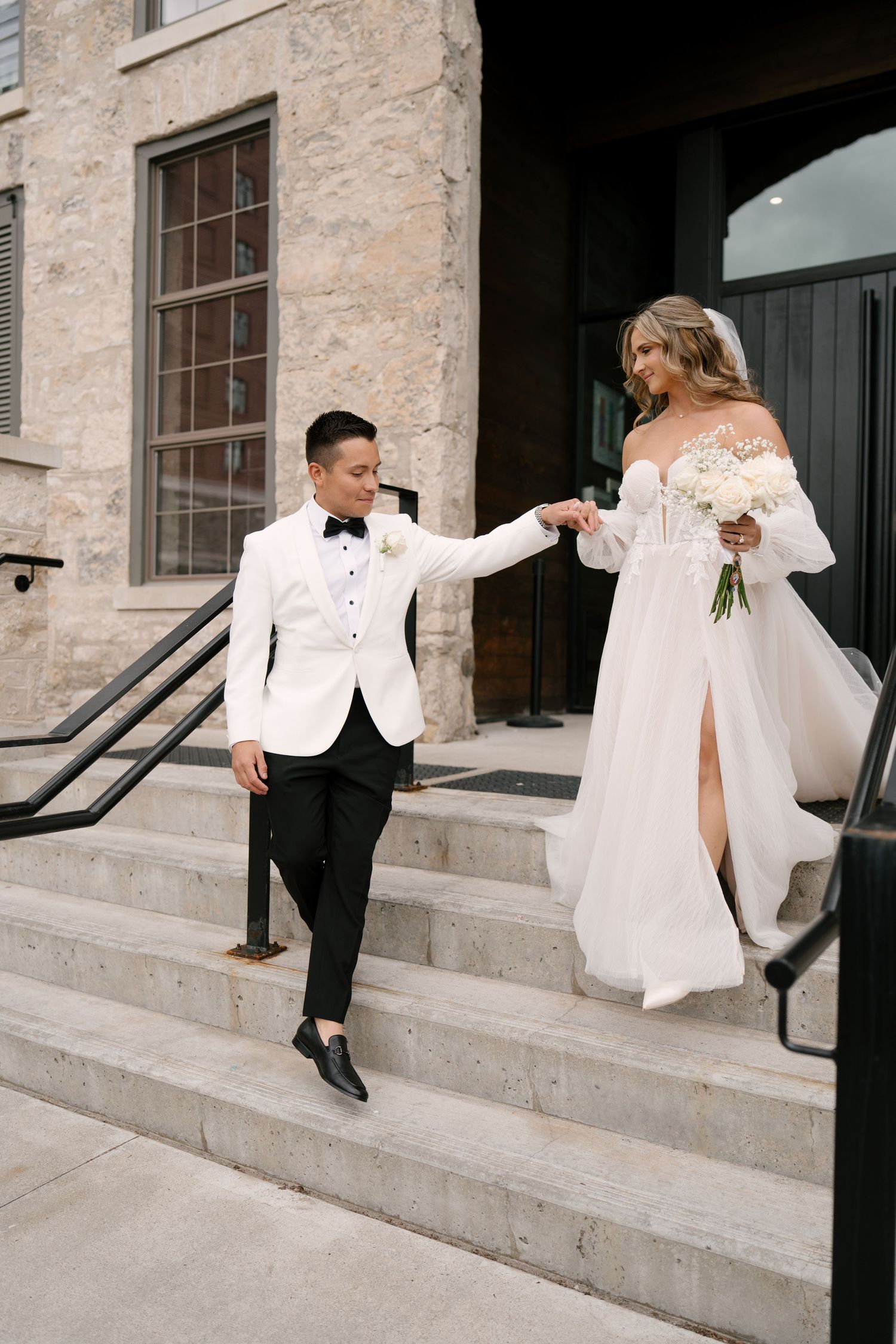 Groom in white jacket leads bride down stone steps while holding her hand at Tapestry Hall venue entrance.