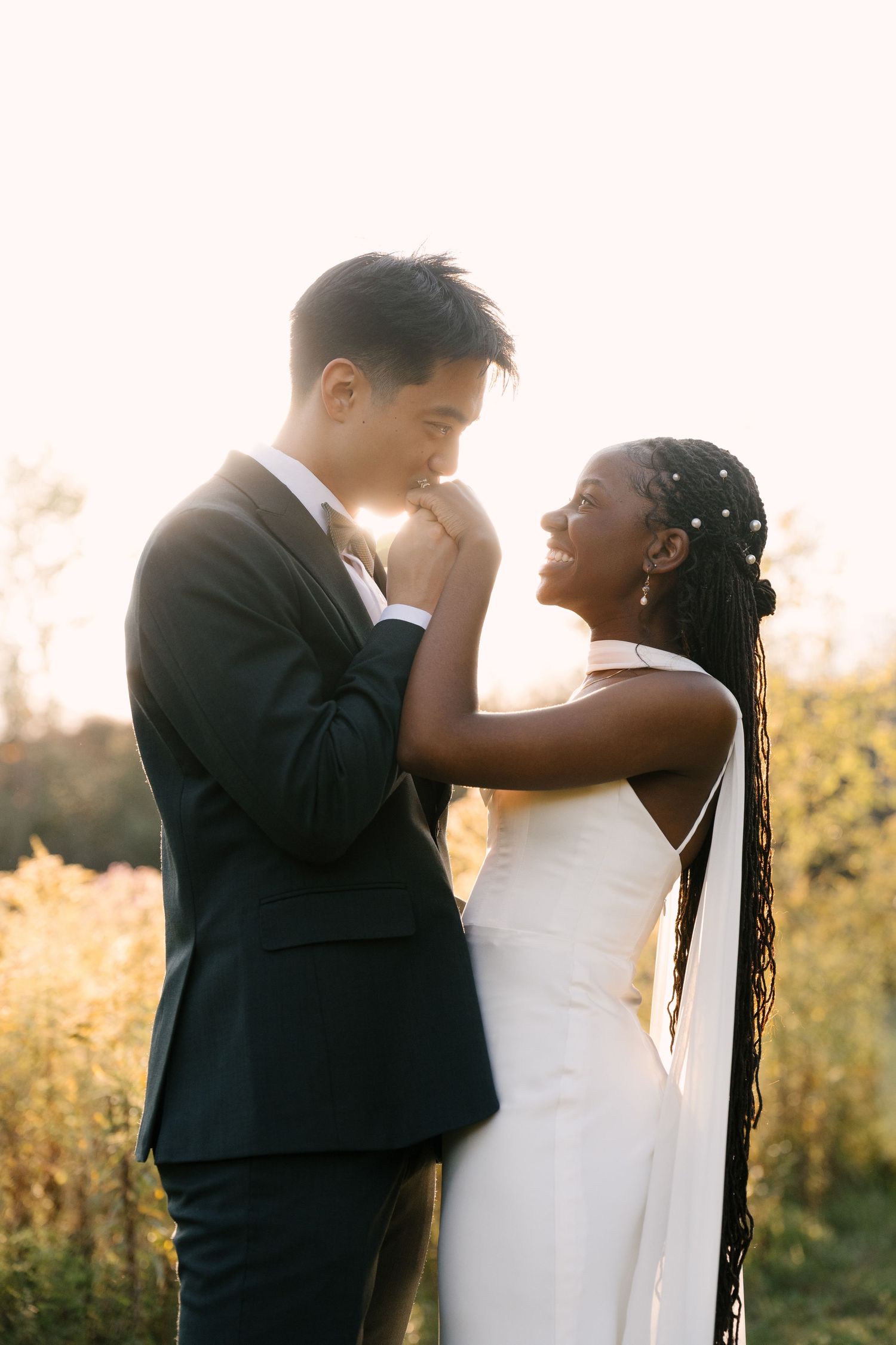 A groom kisses his bride's hand as she smiles joyfully during a golden hour outdoor wedding portrait at Wren House.
