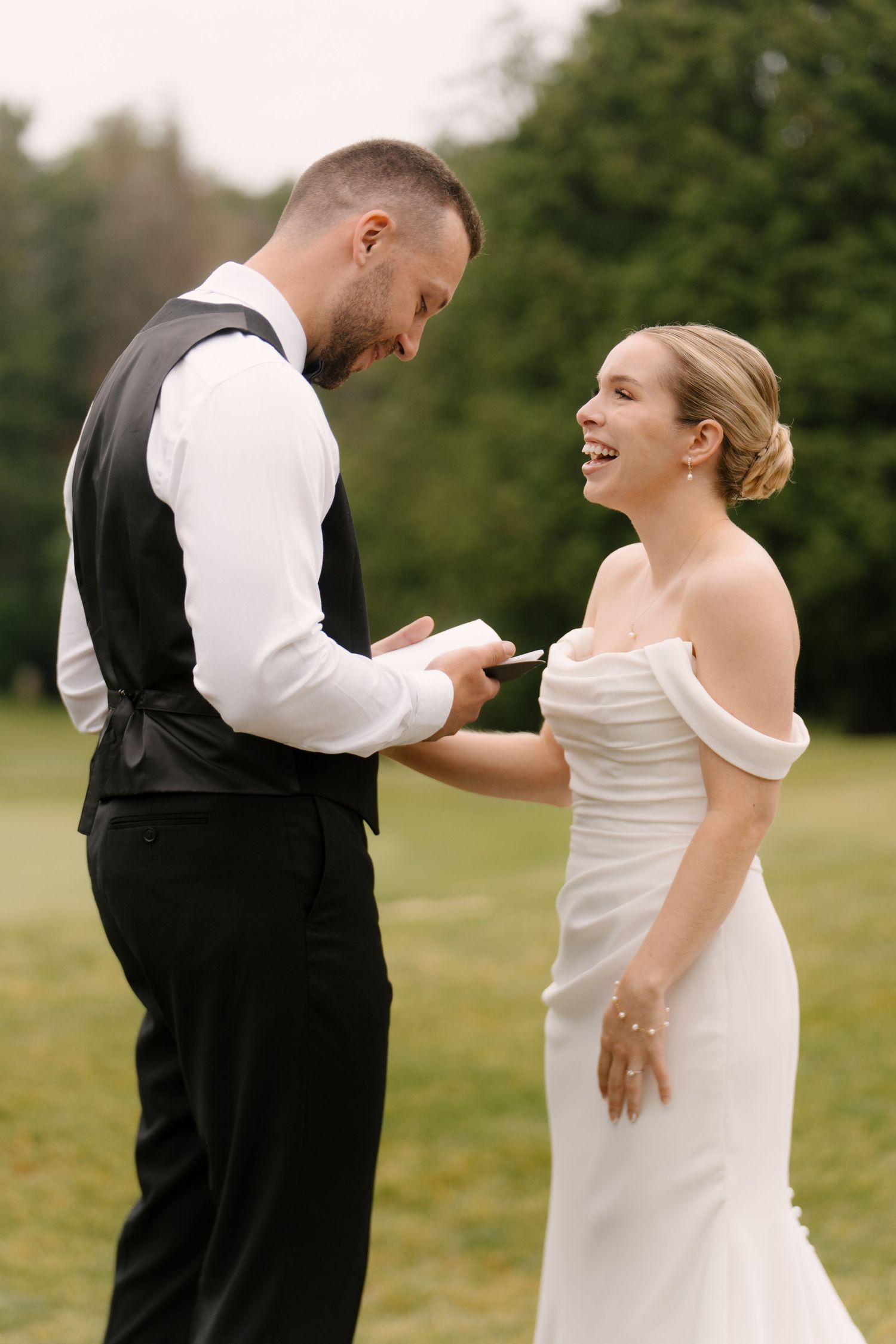 Laughing bride and groom sharing a joyful moment while exchanging handwritten vows outside.