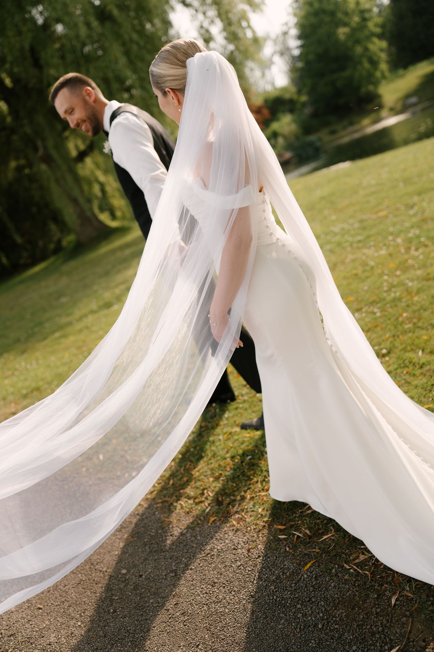 Bride and groom walk a garden path, her long white veil trailing gracefully behind her.