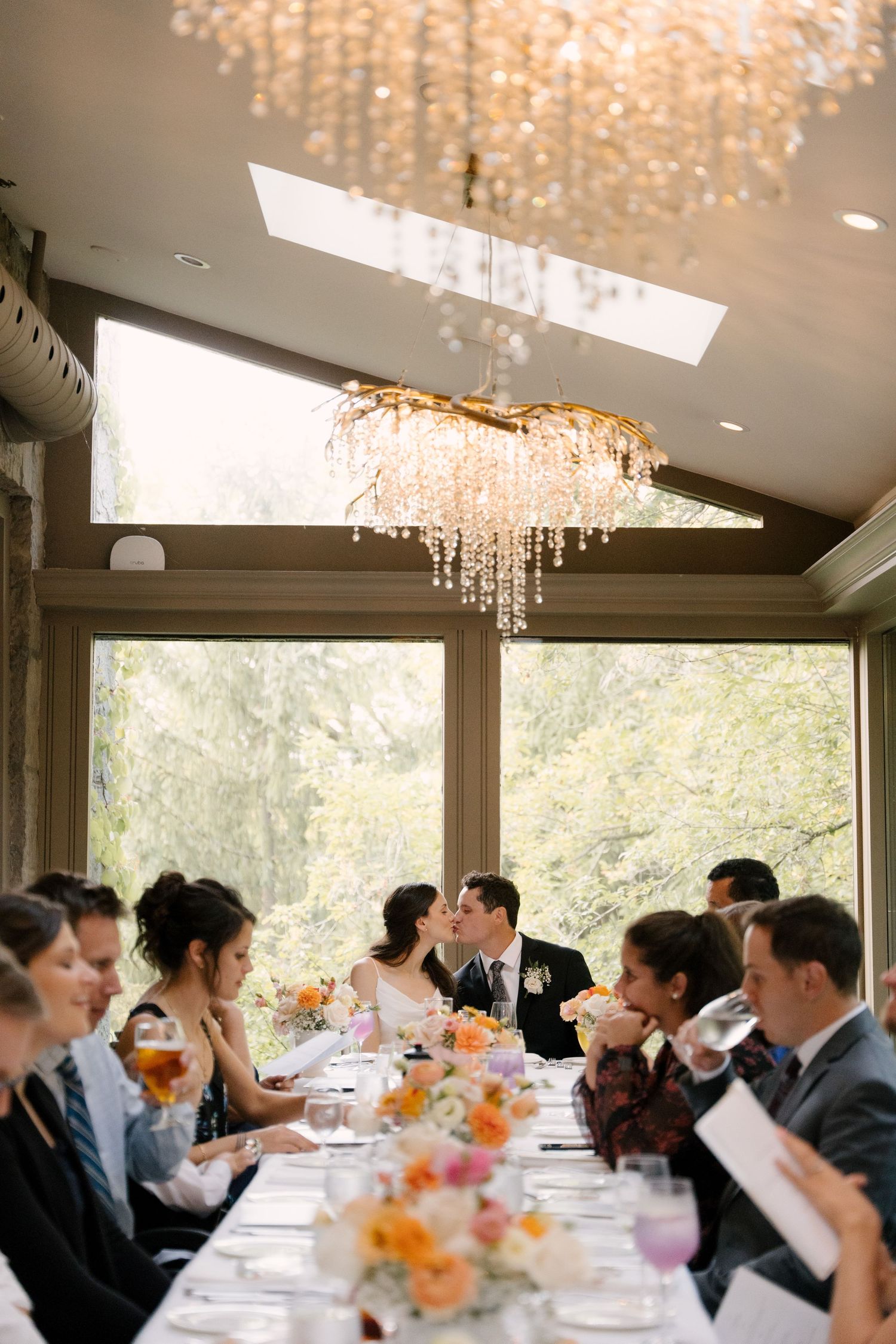 Bride and groom kiss at elegant reception table under crystal chandelier surrounded by guests at Ancaster Mill.