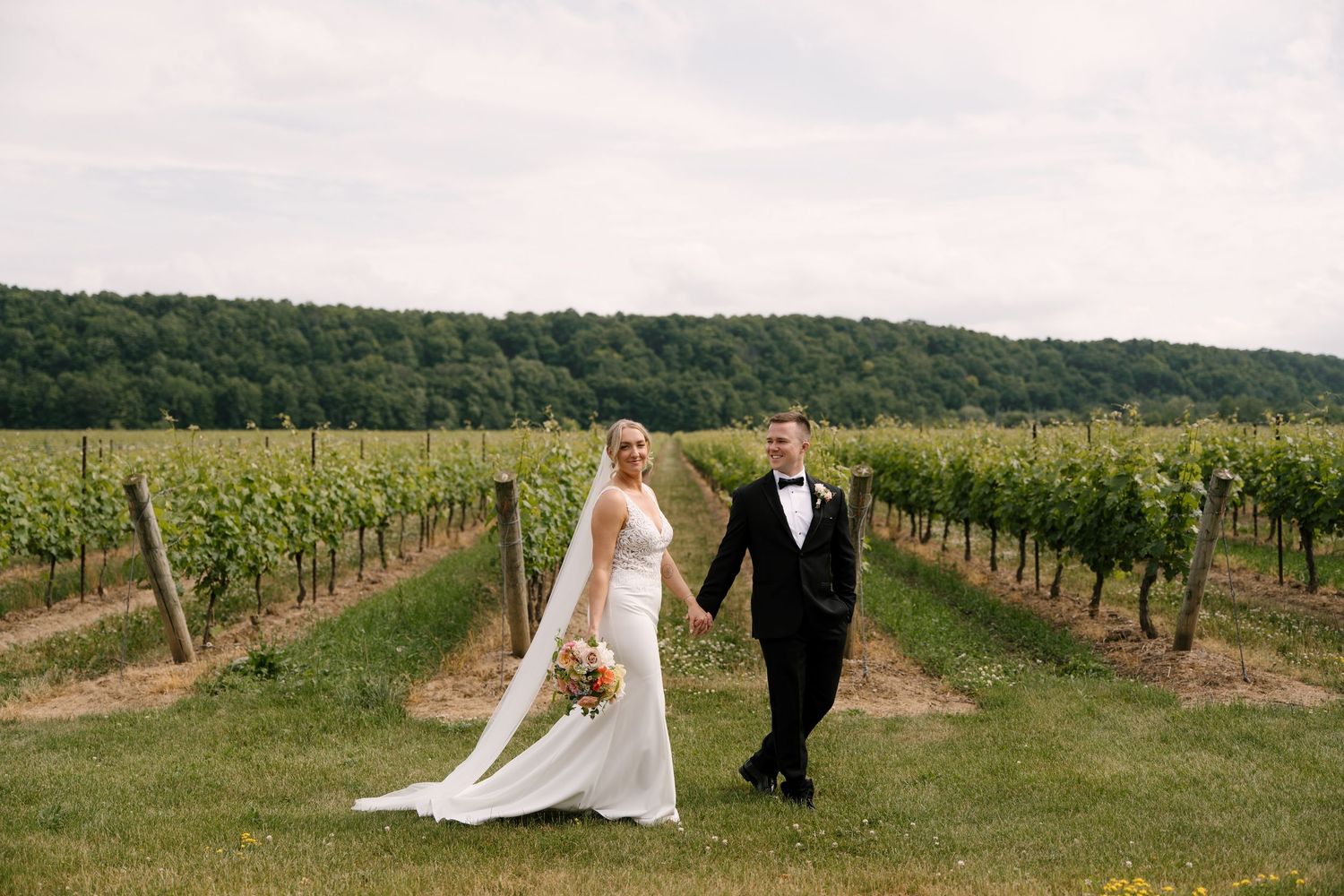 Bride and groom walk hand in hand down center of lush vineyard on wedding day.