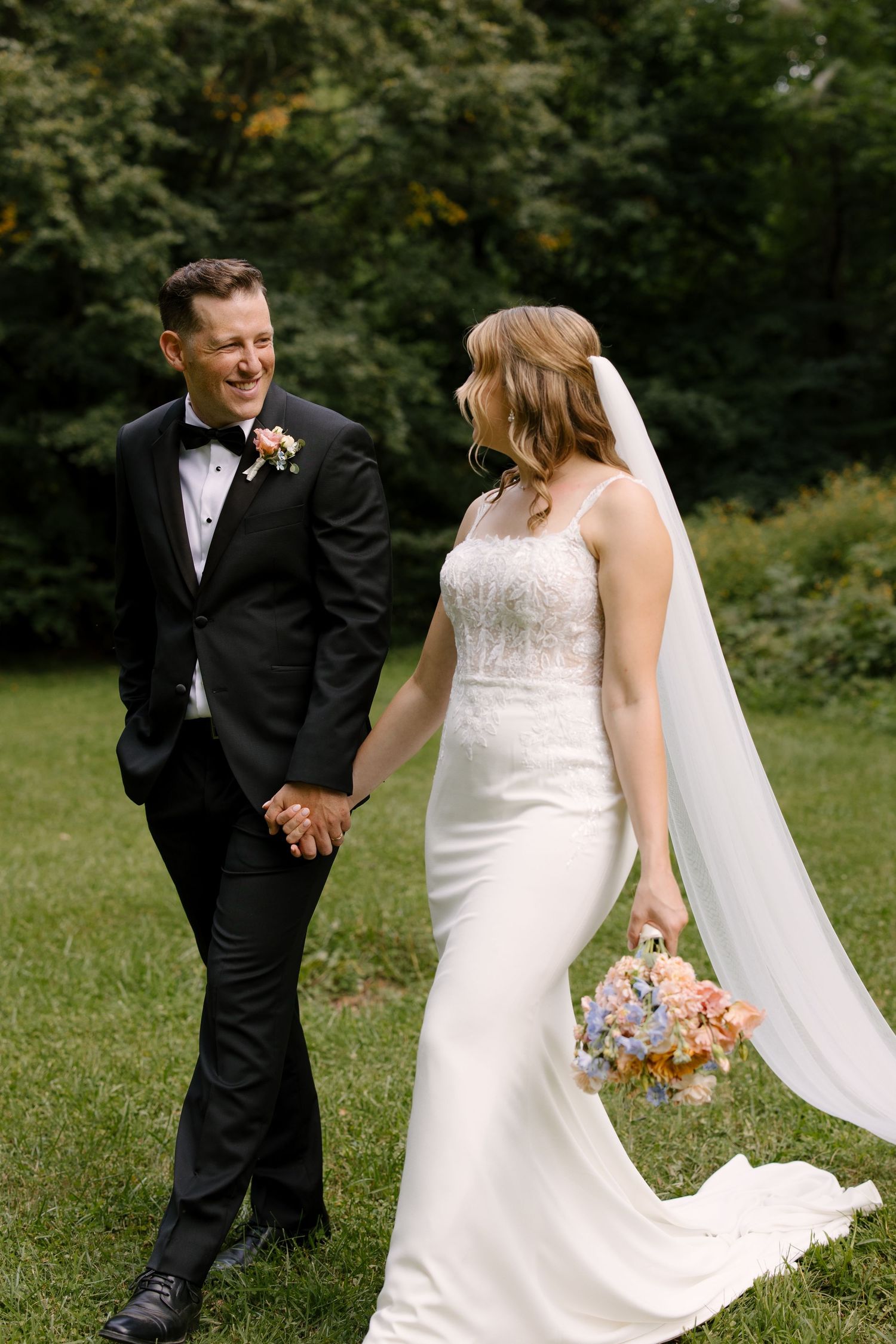 Smiling bride and groom walk hand in hand through green garden, bride holding colorful bouquet.