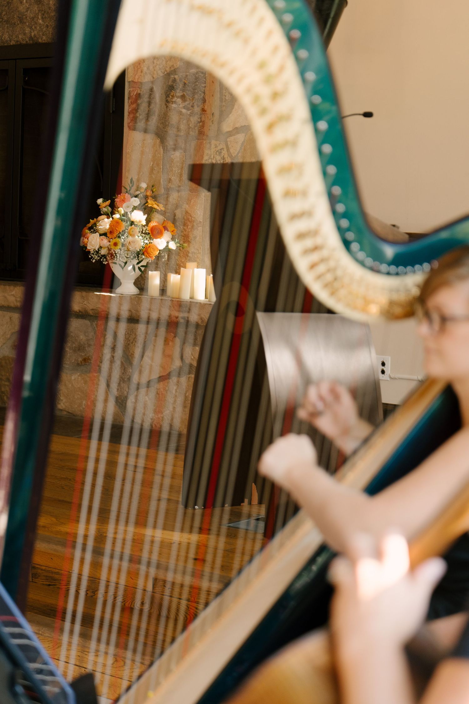 A harpist plays a teal concert harp with floral arrangement and candles reflected in its strings.