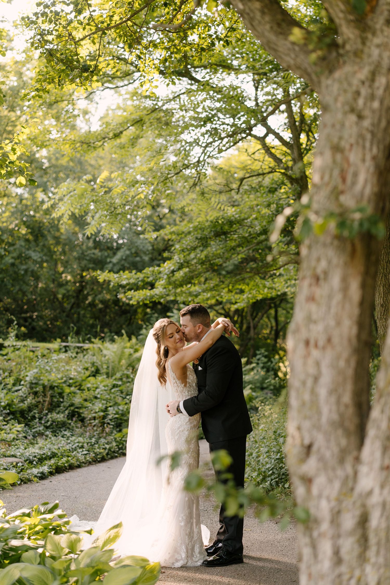 Bride and groom share a romantic embrace on a garden path surrounded by lush green trees and foliage at St Marys Golf Club.