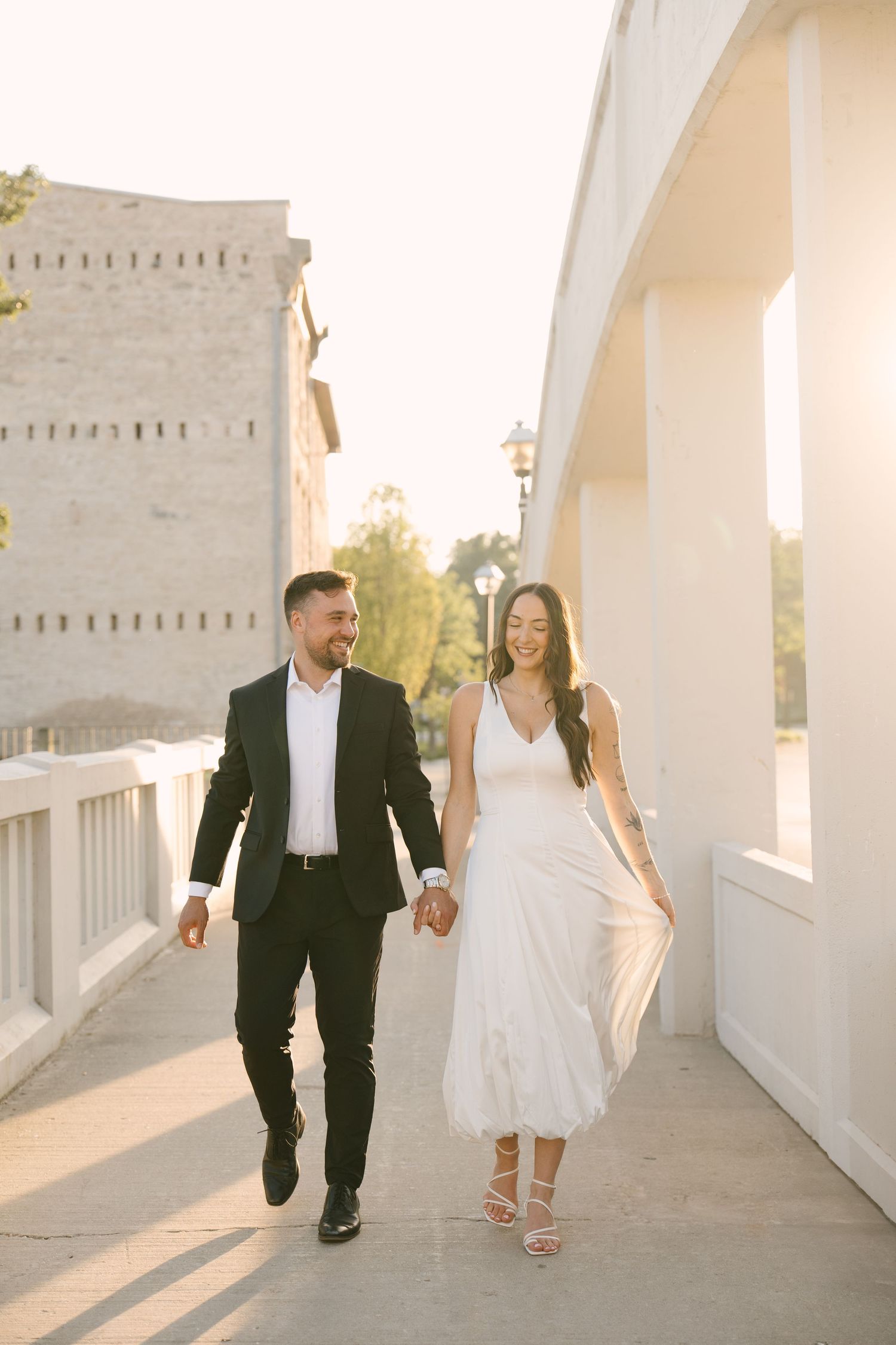 Elegant couple holding hands and walking together along a sunlit outdoor corridor in formal attire.