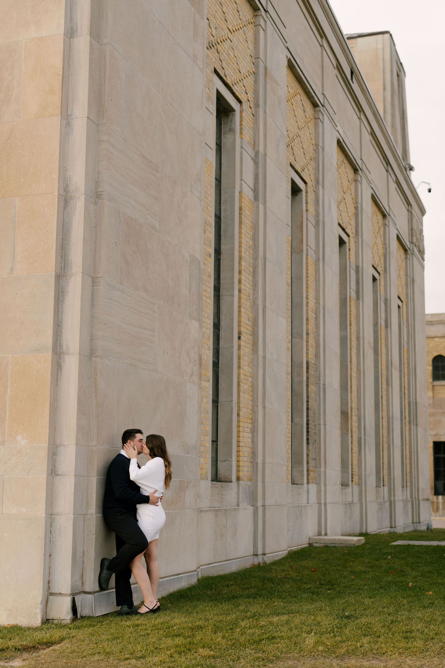 Couple sharing a kiss while leaning against a grand stone building exterior on green lawn.