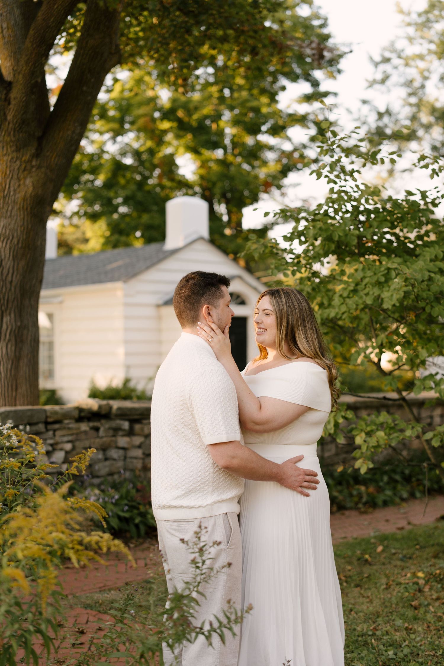 Couple in matching white outfits sharing a tender embrace in a garden with stone wall and cottage backdrop.