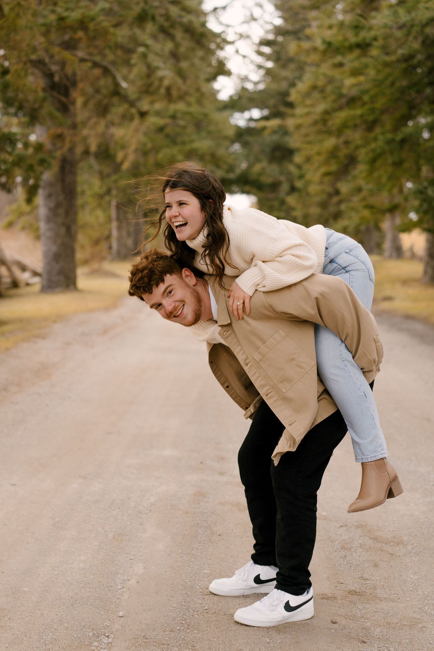 Joyful couple laughing as man gives woman a piggyback ride on a tree-lined gravel road.