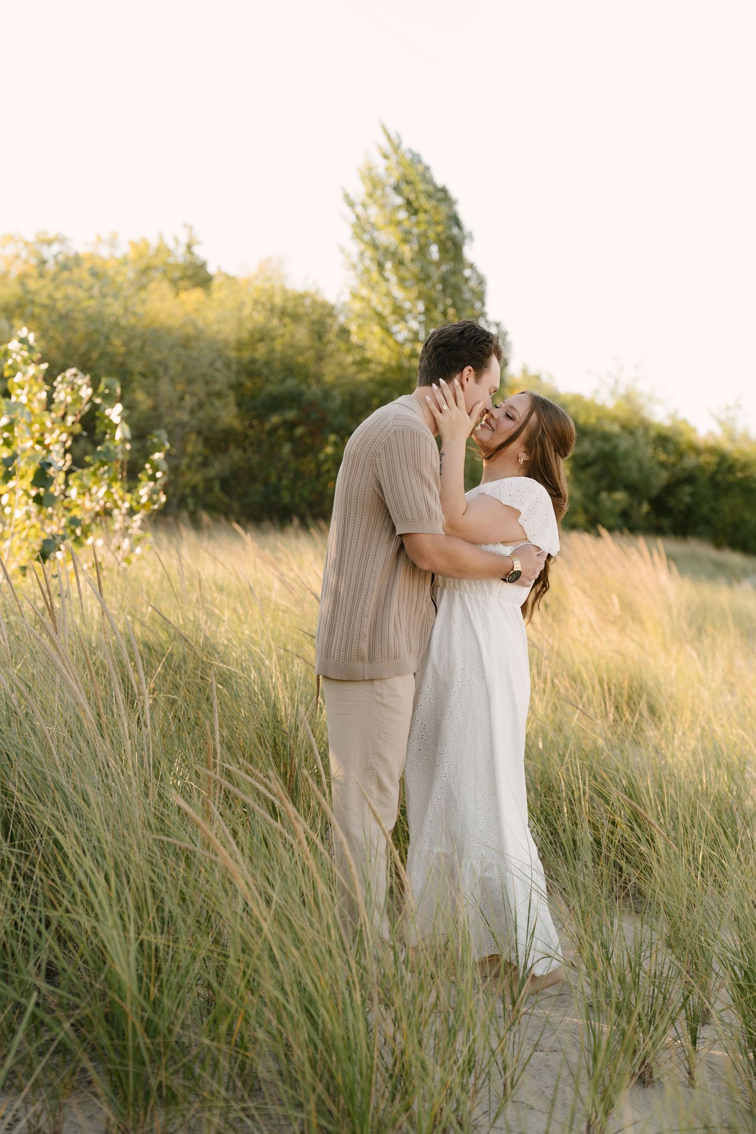 Couple sharing a kiss among tall beach grass at golden hour sunset.