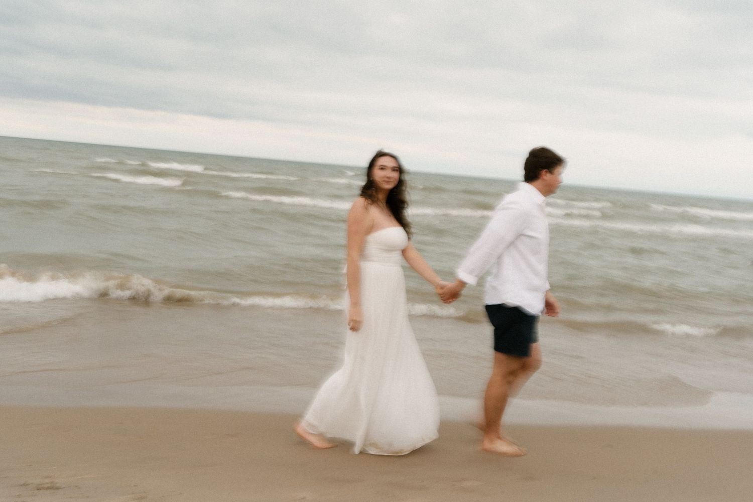 Couple walking hand in hand along ocean shoreline, woman in elegant white strapless gown, man in white shirt.