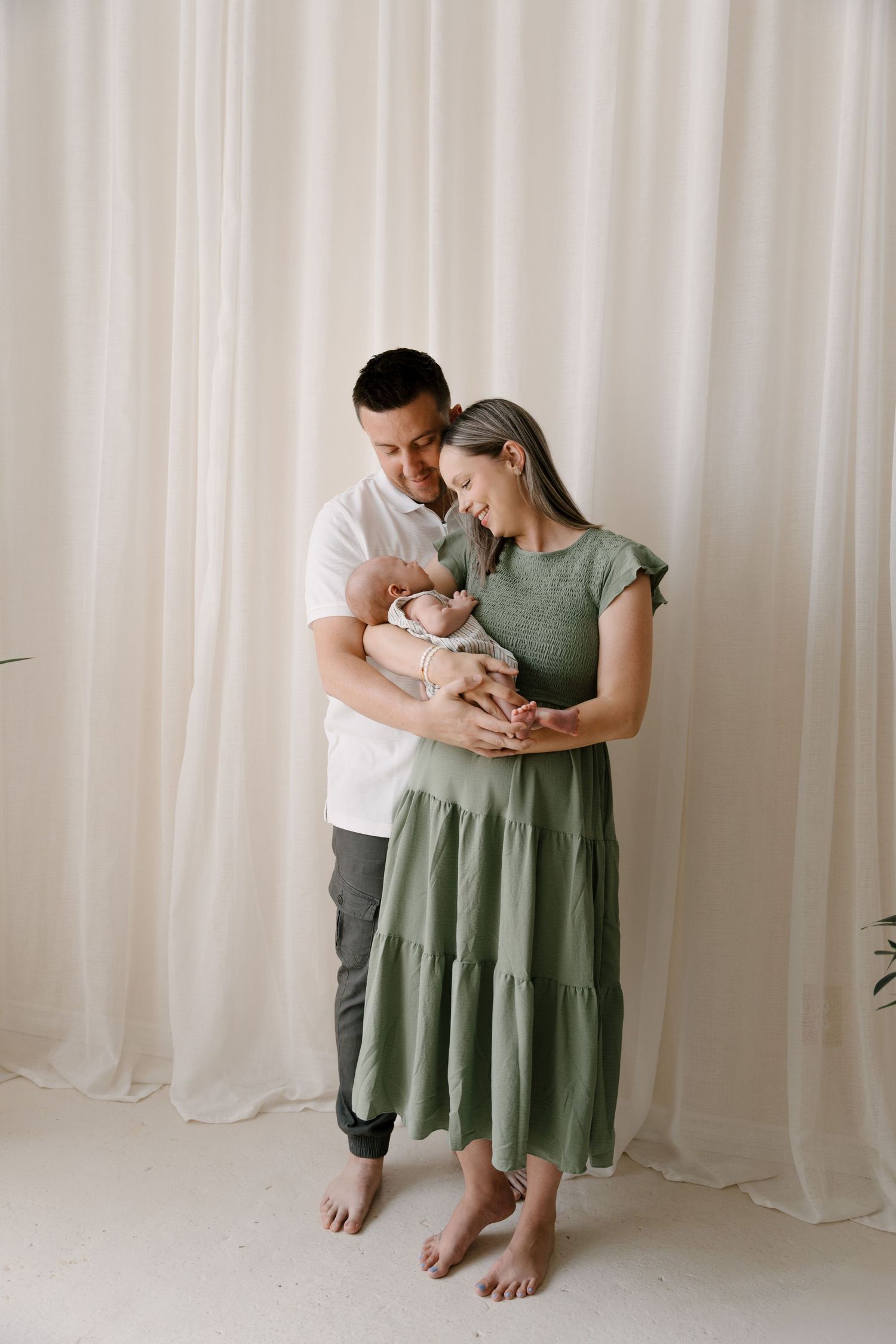 A smiling couple in a bright studio holds their newborn baby, mother wearing a green tiered dress.
