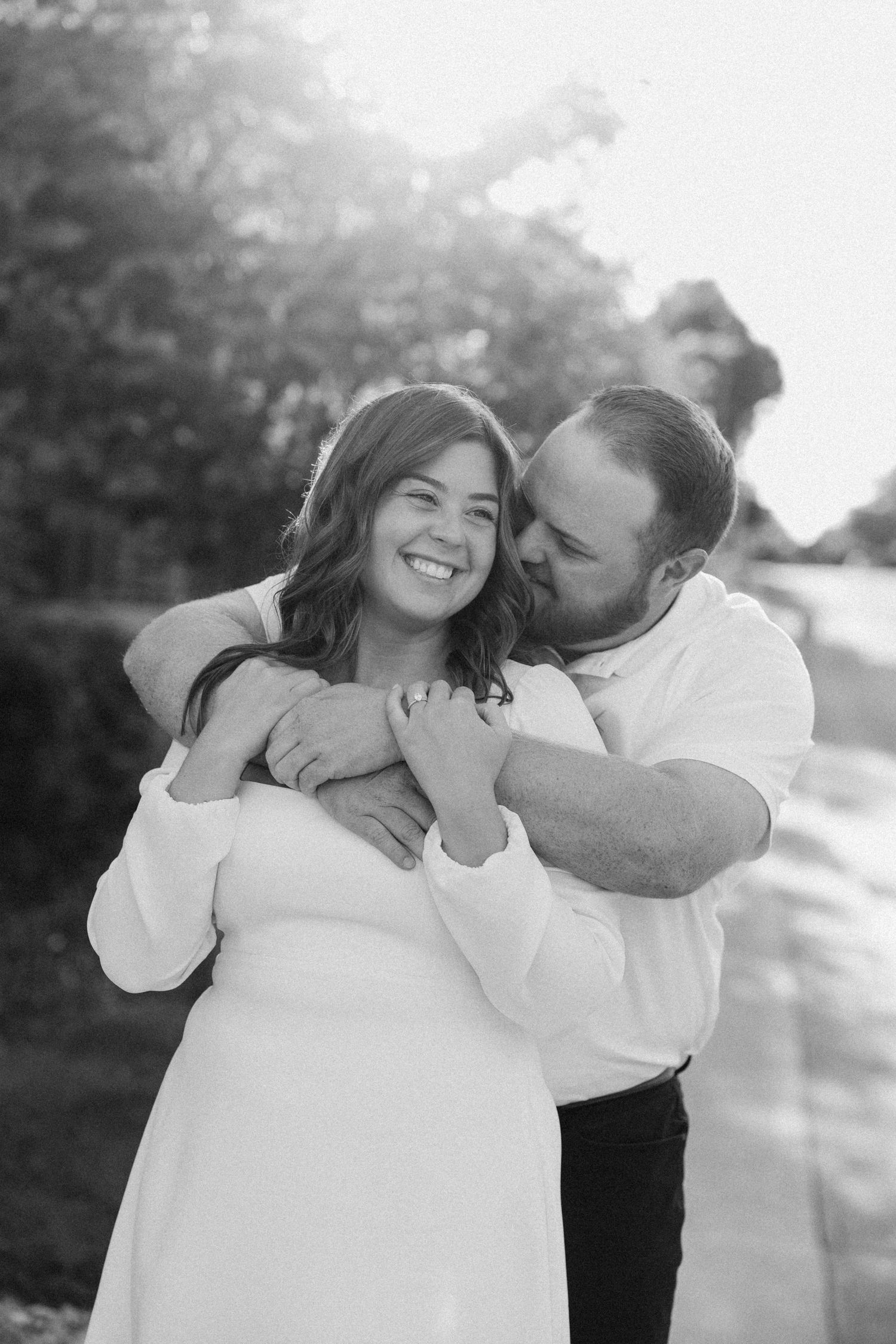 Black and white photo of man embracing woman from behind as she smiles joyfully outdoors.