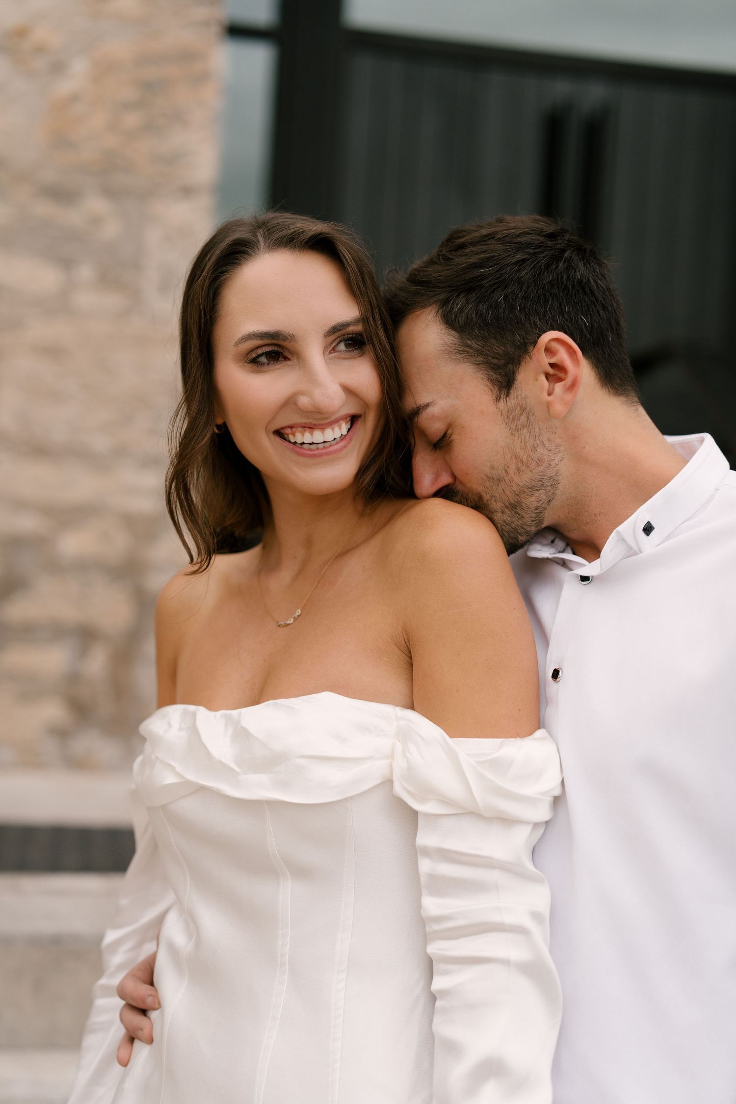 Woman in white off-shoulder dress smiling as man kisses her shoulder outdoors.