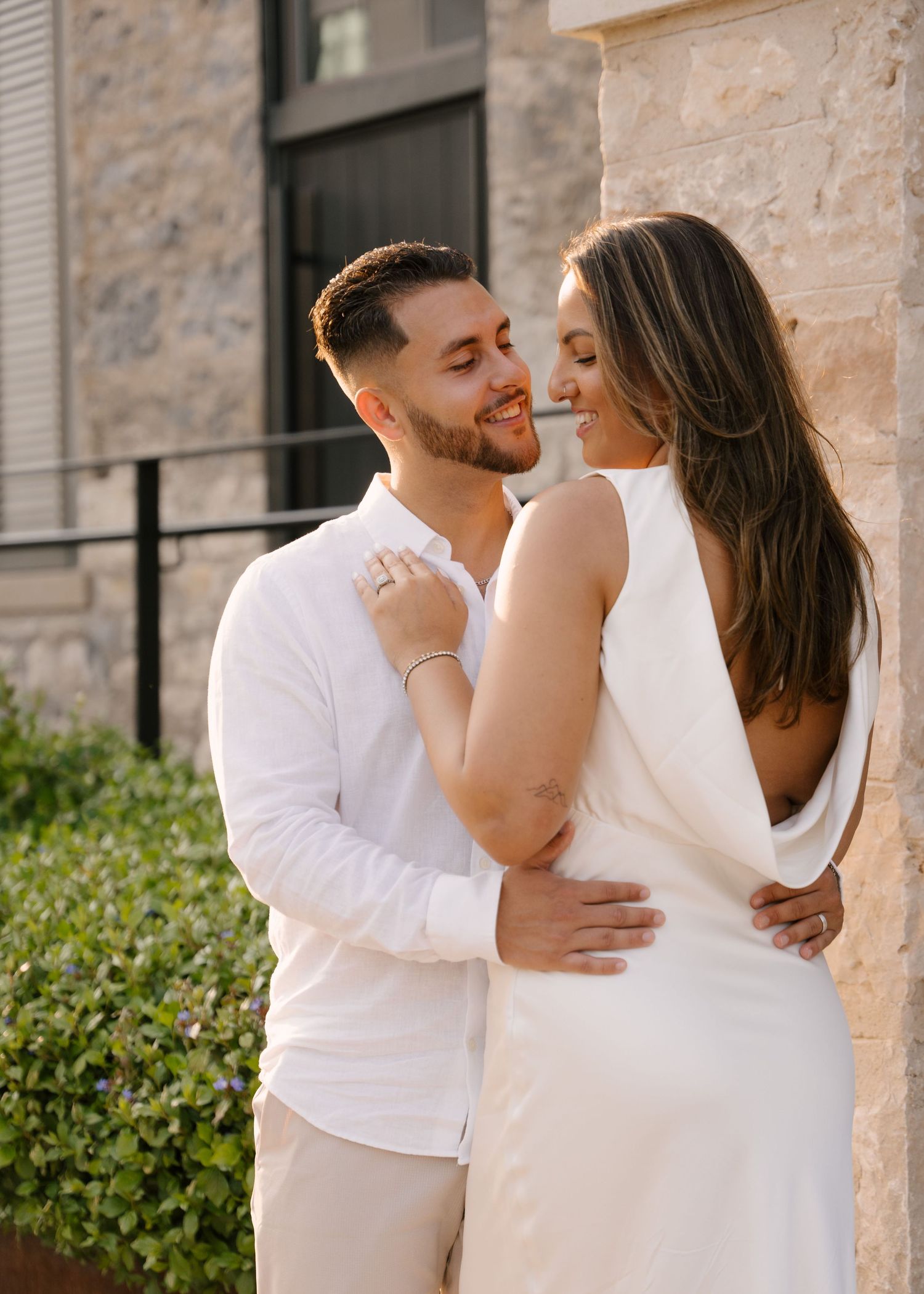 A smiling bearded man embraces a woman in a backless white dress against a stone wall outdoors.