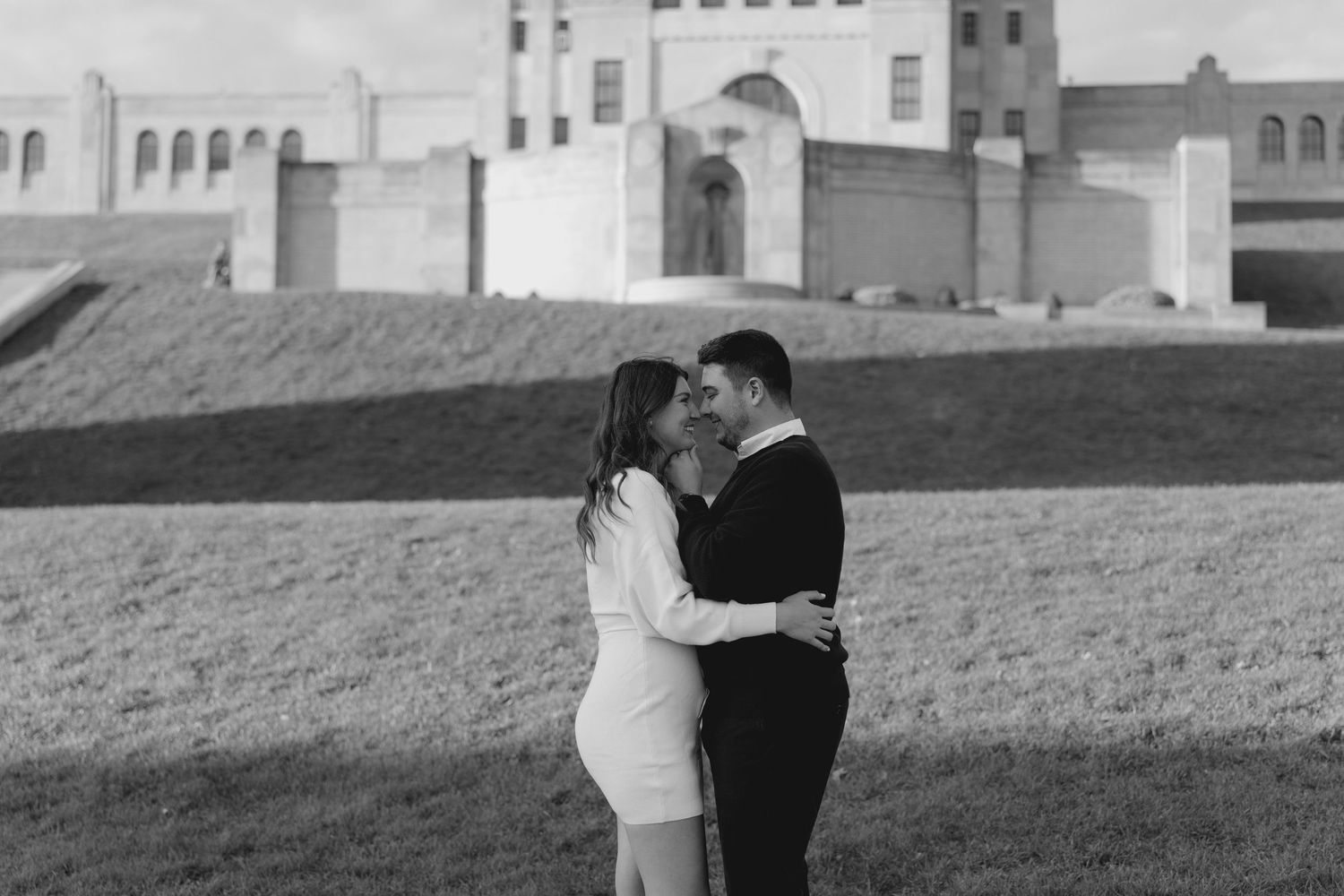 Black and white photo of couple sharing a kiss near historic stone building on open green lawn.