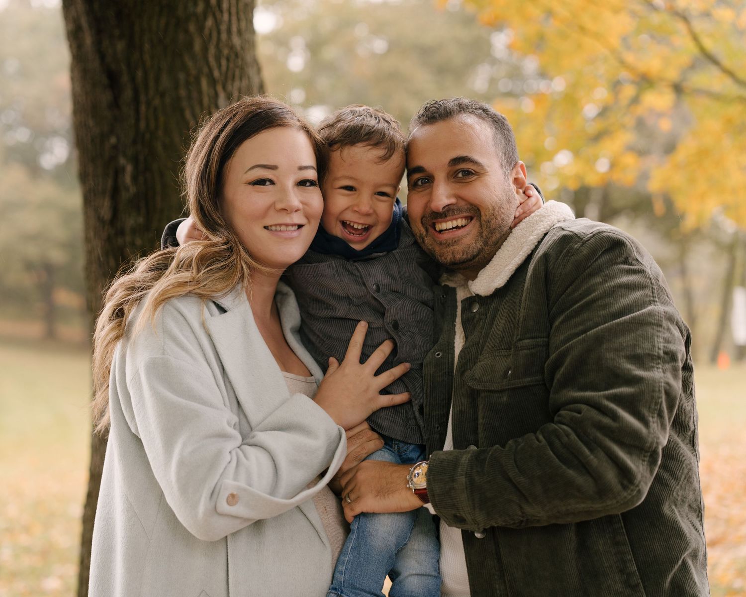 Joyful family portrait with pregnant mother, father, and toddler son among fall foliage in park.
