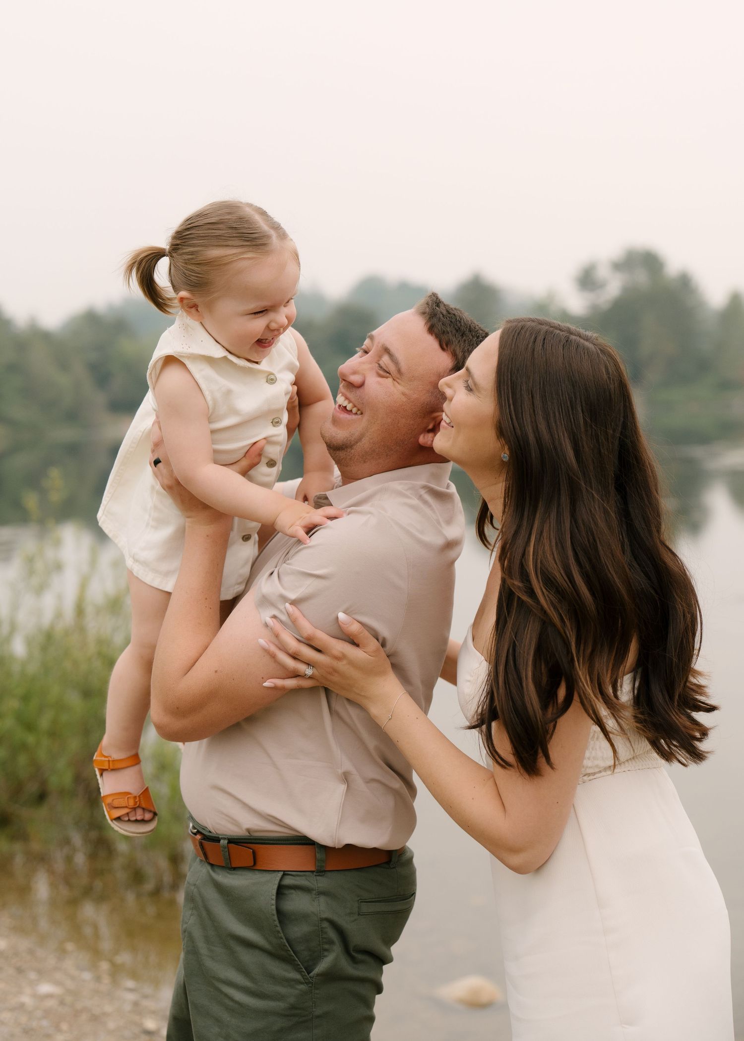 Happy family portrait with parents laughing and holding toddler daughter by the water.