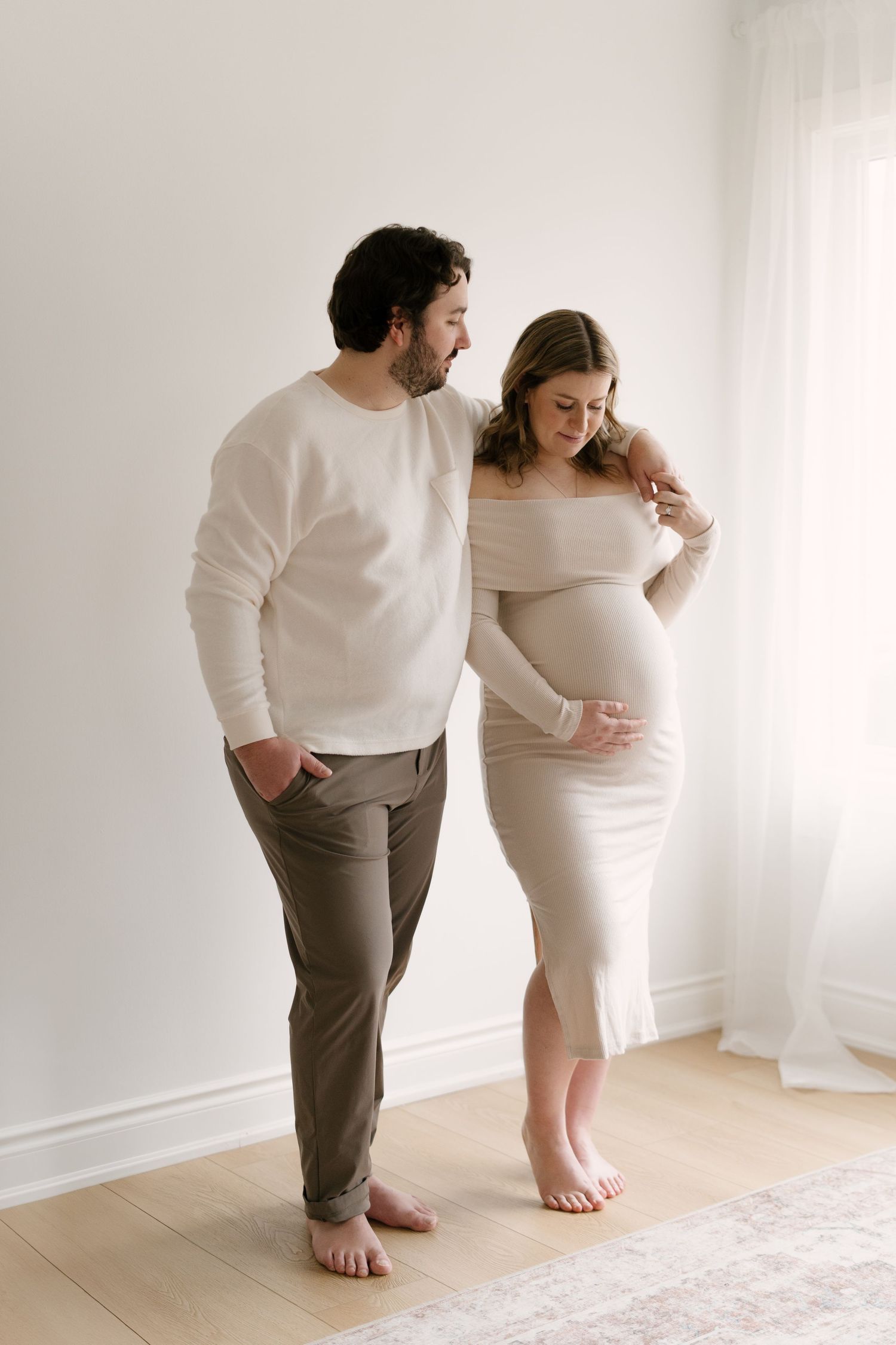 Expecting couple in neutral tones standing together, man gazing at pregnant woman holding her baby bump.