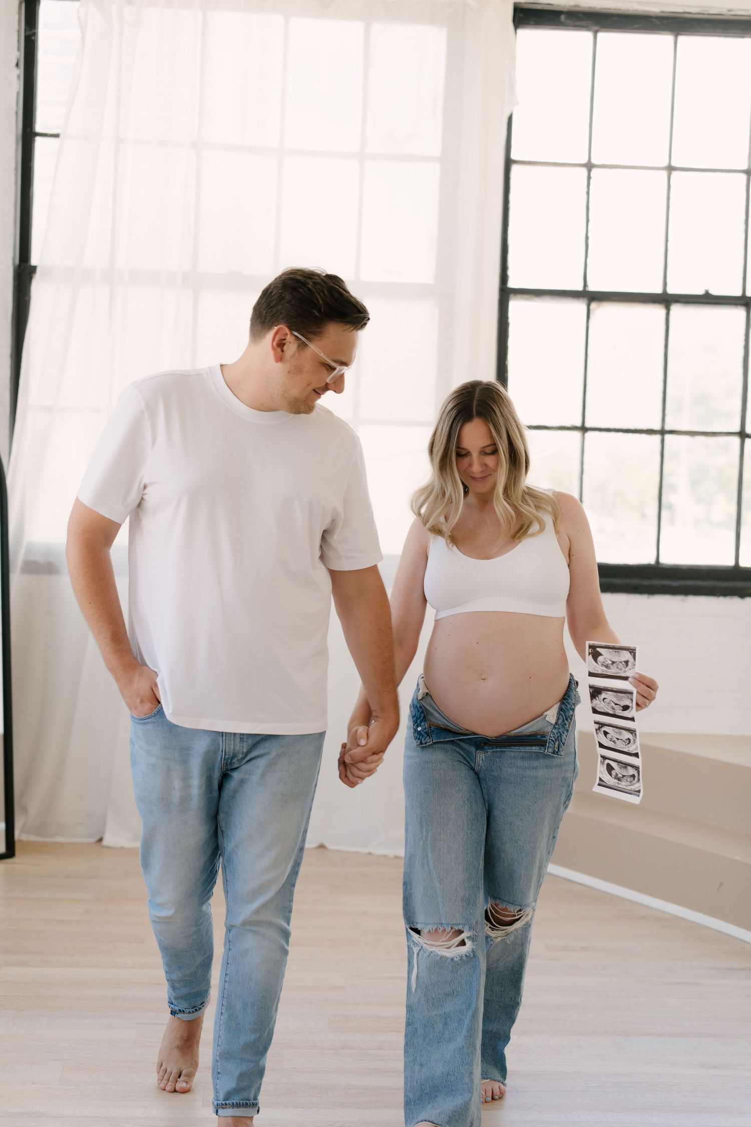 Couple holding hands and looking down at ultrasound photos held by pregnant woman in white crop top.
