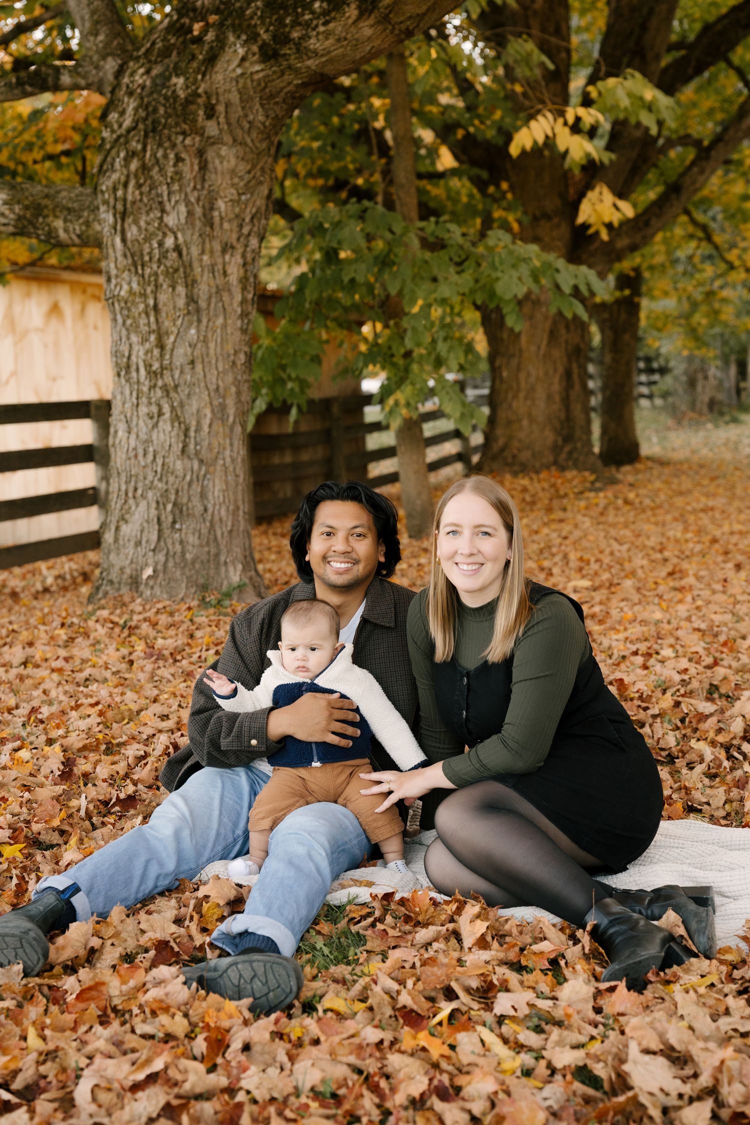 Interracial family sitting together on fall leaves under large trees, holding baby.